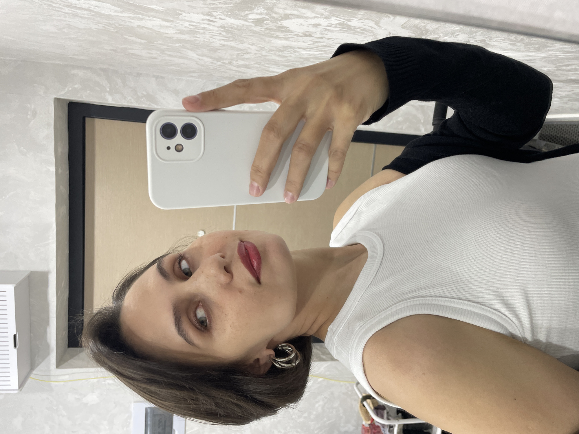 A woman is taking a selfie. She is holding a white smartphone with her left hand. She is wearing a white tank top and a black sleeve. She has short brown hair and is wearing silver hoop earrings. Her lips are painted red. The background includes a light-colored wall and a door frame. The location is not identifiable as Izhevsk, Russia.