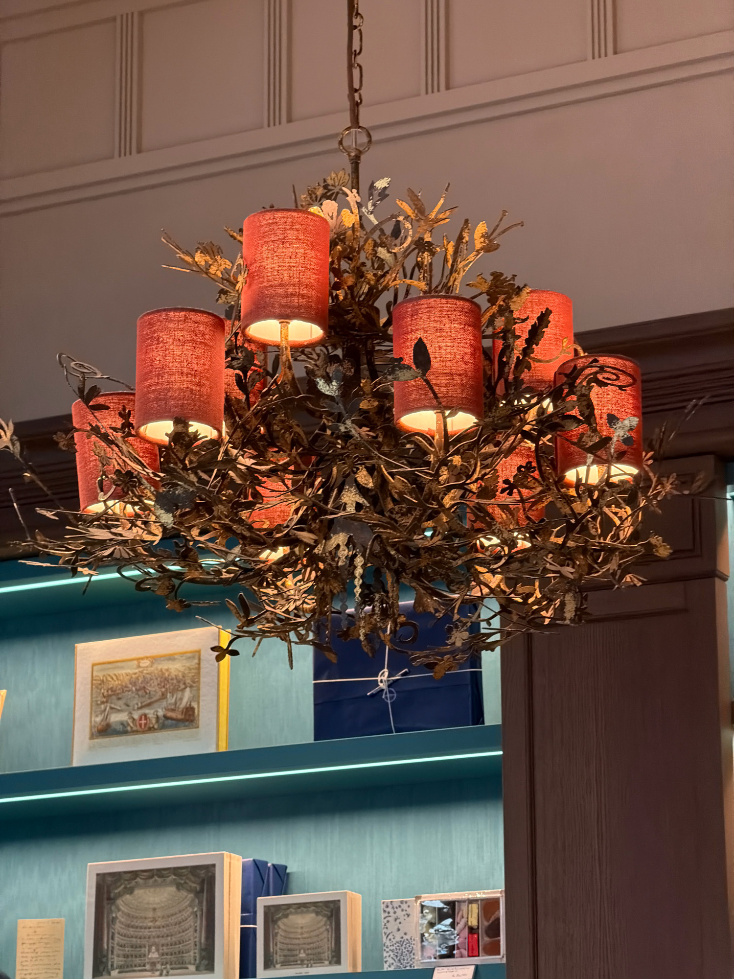 The image shows a close-up of a decorative chandelier with red drum-shaped lampshades hanging from a chain. The chandelier is ornate, with metal branches and leaves resembling a vine. Below the chandelier, on teal shelves, are framed pictures and boxes. One framed picture shows a historical city scene with ships, another displays an interior of a theater or opera house, and a smaller framed picture shows a similar theater interior. Several blue wrapped boxes are also visible, along with a small open box containing what appear to be candies or pastries. The overall setting suggests a retail or display environment. This scene is located in Milan, Italy, specifically within the historic La Scala opera house.