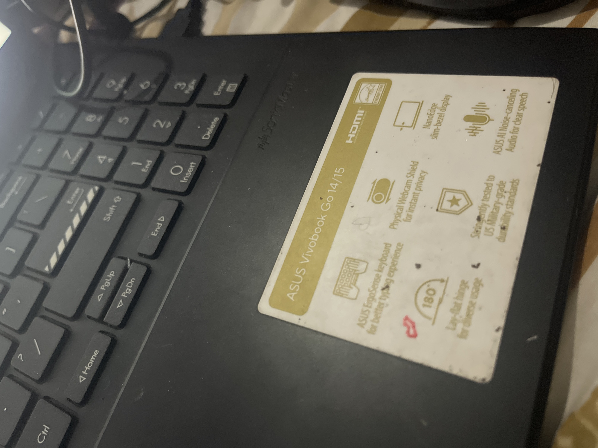 The image shows a close-up of a black ASUS Vivobook Go 14/15 laptop keyboard and a sticker on the palm rest. The keyboard has standard QWERTY layout keys, including "Ctrl," "Home," "PgUp," "PgDn," "End," and "Insert." The sticker features the ASUS Vivobook Go 14/15 branding and highlights various features like the ASUS ErgoSense keyboard, a physical webcam shield, a lay-flat hinge, NanoEdge slim-bezel display, and ASUS AI Noise-canceling audio. The "HDMI" logo and "ENERGY STAR" certification are also visible on the sticker. The laptop appears to be placed on a surface with a patterned cloth, possibly a blanket or tablecloth, suggesting an indoor environment. The lighting is dim, and the image has a slightly blurry quality. There are no people or specific Santa Ana, Venezuela landmarks visible in this image.