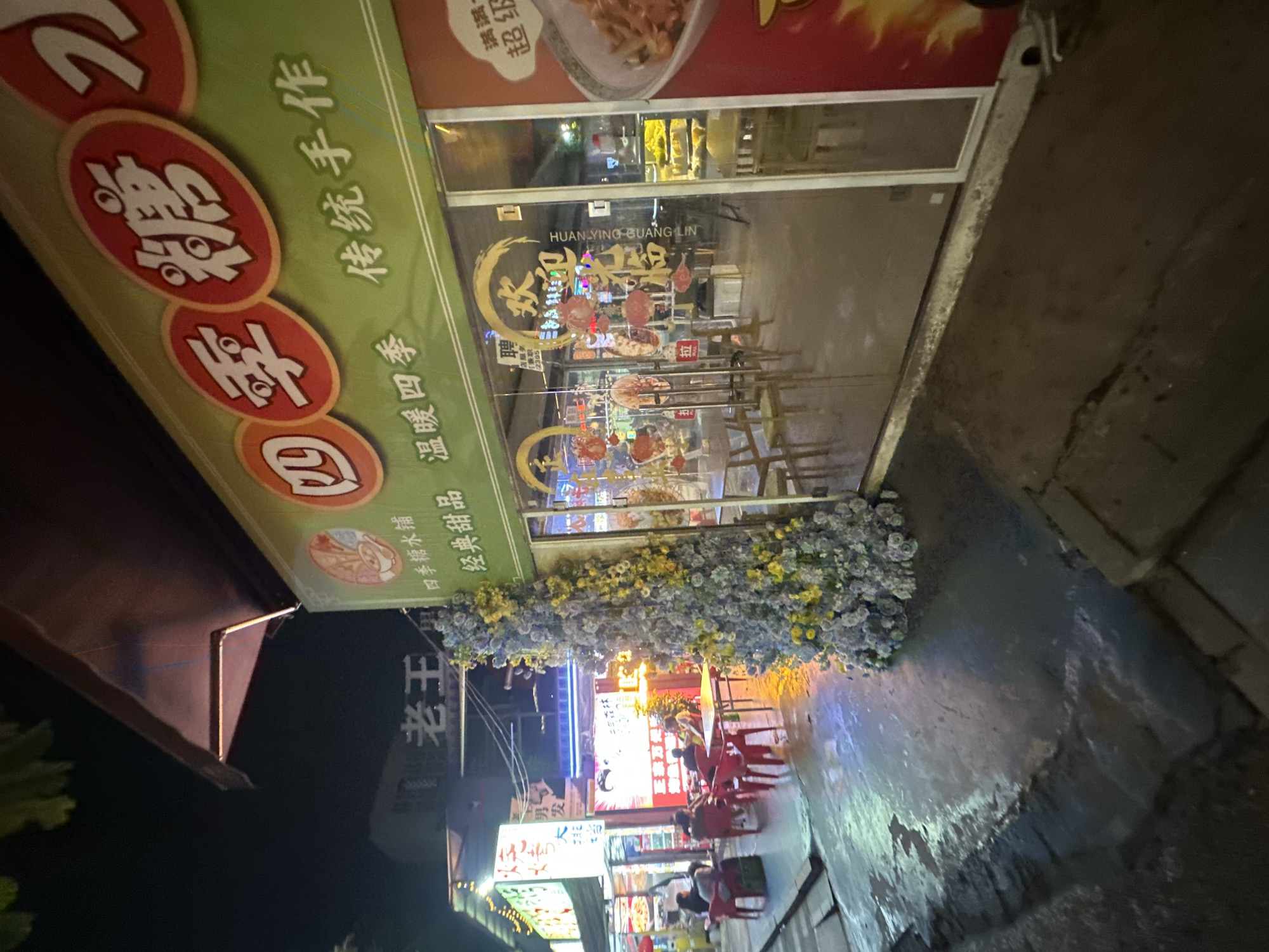 The image depicts a nighttime scene on a street, possibly in the Chongqing High-tech Zone, China. The focal point is the storefront of a dessert shop with bright green signage featuring Chinese characters and circular emblems. The storefront has large glass windows revealing the interior, where tables and chairs are visible, along with illuminated displays. Outside the shop, the wet pavement reflects the colorful lights from the establishment and surrounding signs, indicating recent rain or street cleaning.
To the left of the dessert shop, another storefront with illuminated signage is partially visible, and several people are seated at tables under a canopy. A tall, densely decorated floral arrangement, primarily in shades of blue and yellow, stands near the entrance of the dessert shop. The overall atmosphere is one of a bustling night market or commercial street.