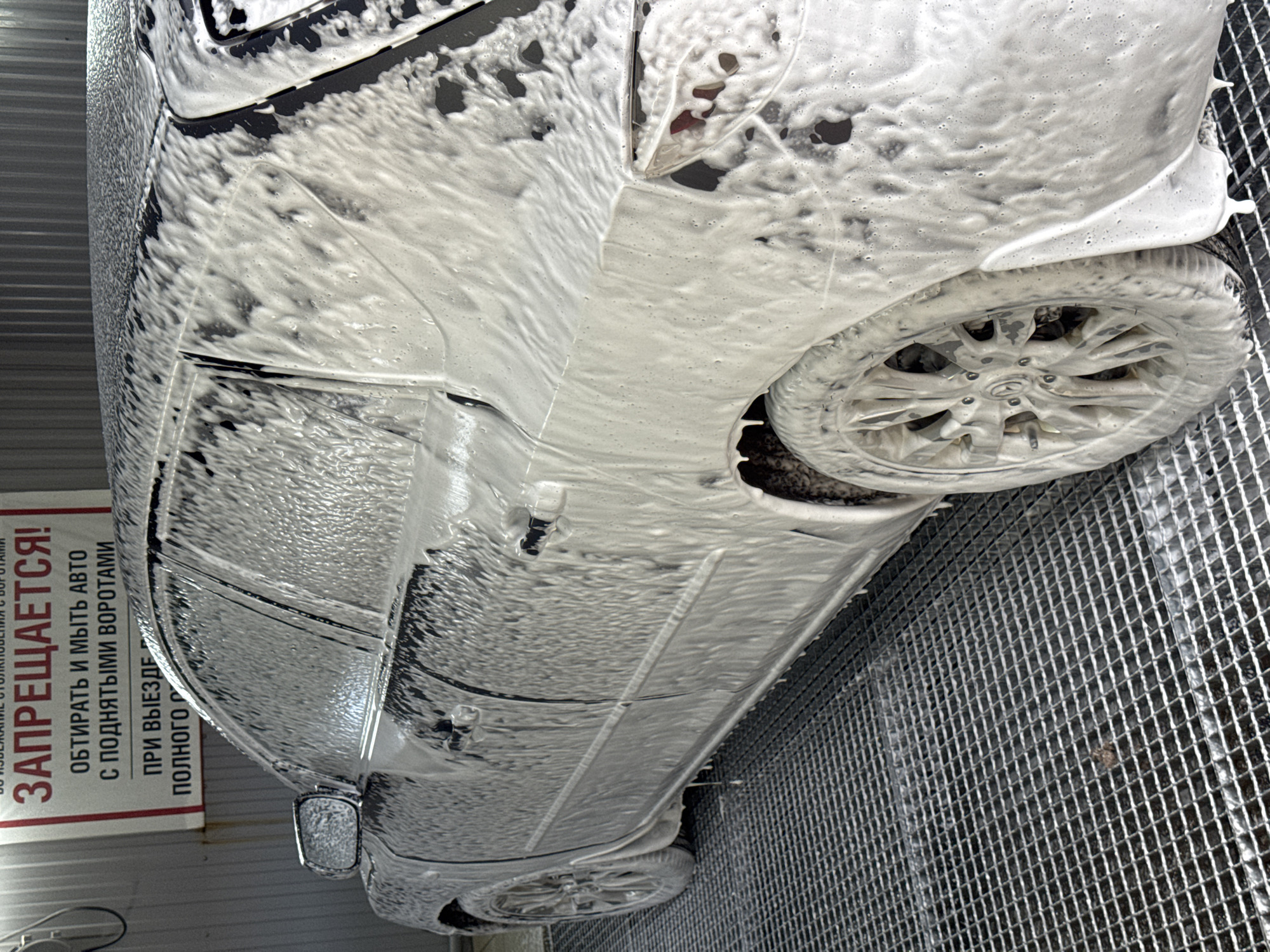 The image shows a dark-colored car covered in soap suds, undergoing a car wash in Chaikovsky, Russia. The car is positioned on a metal grate floor. A sign in Russian is visible, prohibiting the wiping and washing of cars with the gate open until completely off the property. The car's wheel is also covered in soap. The environment is a car wash facility.