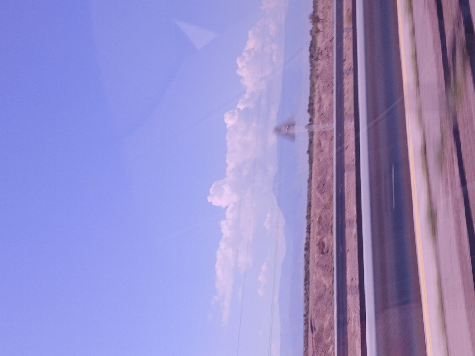 This is a landscape photograph taken from a moving vehicle, likely on a highway. The sky is a pale blue with some wispy clouds, and the horizon is dominated by a range of distant, hazy mountains. In the foreground, a multi-lane highway is visible, with guardrails and clear lane markings. The landscape flanking the highway appears arid and sparsely vegetated, with low hills and some scattered trees. A blurred traffic sign is visible on the left side of the road. The overall impression is one of a vast, open, and somewhat desolate environment. The location in the city of Nurly, Kazakhstan, is not explicitly indicated by any visual cues within the image itself.