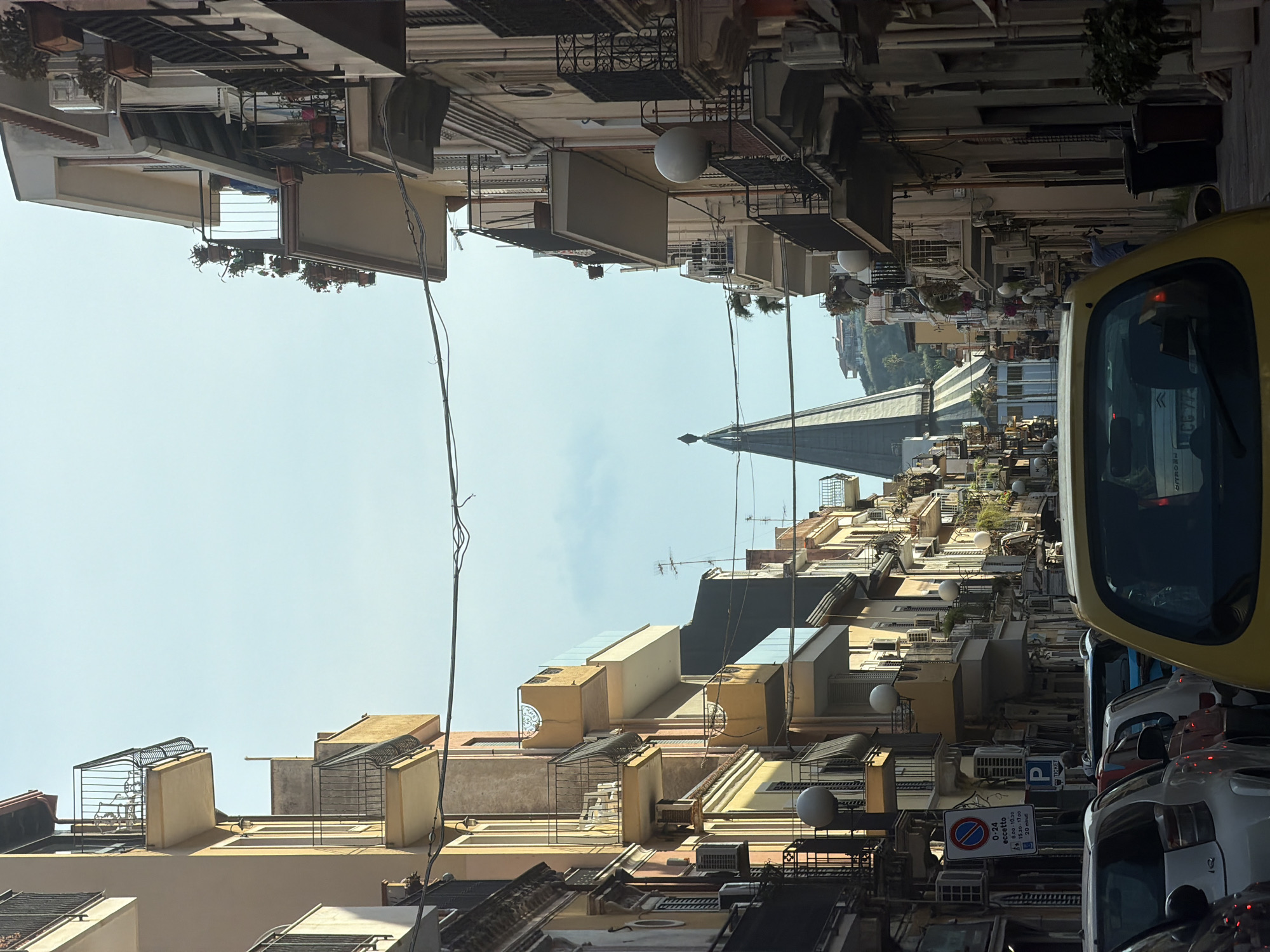 The image depicts a narrow street in Chianchitta, Italy, densely lined with multi-story buildings. The perspective is tilted upwards, emphasizing the verticality of the architecture. The buildings are a mix of pale yellow, beige, and light brown hues, featuring balconies, air conditioning units, and various architectural protrusions. The sky is visible between the buildings, with a tall spire or tower-like structure partially visible in the distance. The street is congested with parked and moving vehicles, including a yellow car prominent in the foreground. A blue and white parking sign is visible on one of the buildings. There is a sense of urban density and the composition suggests a cramped, bustling environment. No people are clearly visible in the scene.