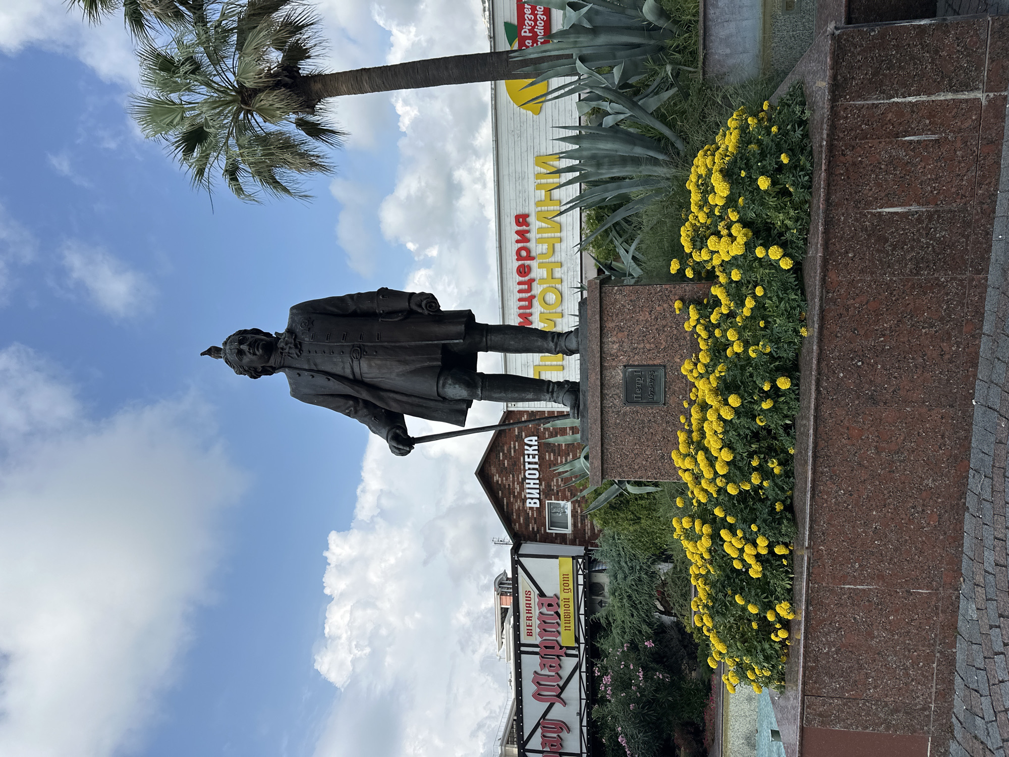 The image shows a bronze statue of a man, possibly Peter the Great based on the inscription on the pedestal, standing on a granite base surrounded by yellow marigolds. The statue is located in Sochi, Russia, as indicated by the inscription on the pedestal and the presence of palm trees and agave plants which are common in the region. In the background, there are several buildings with signs in Russian, including "Виннотека" (Vinoteka - wine shop) and "Mary Marma" with "BIERHAUS" and "пивной дом" (pivnoy dom - beer house). There is also a sign for "ЛИМОНЧИНИ" (Limochini - lemon trees), which appears to be a restaurant or cafe. The sky is partly cloudy with some white cumulus clouds. A small bird is perched on the head of the statue. The overall scene depicts an outdoor public space with a monument and commercial establishments.