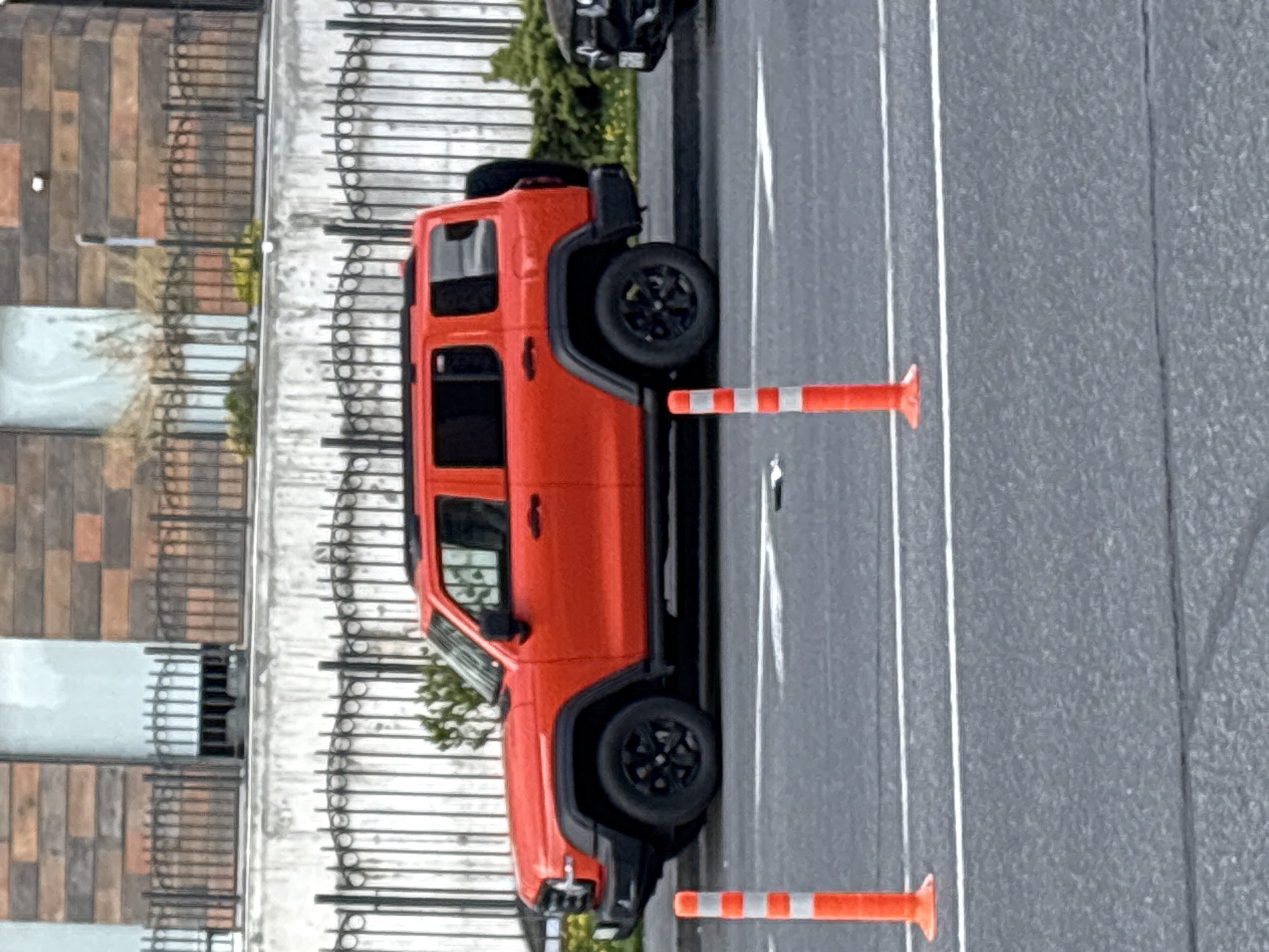 An orange four-door Jeep Wrangler is prominently featured, parked on a grey paved surface that appears to be a street or parking lot. The vehicle is oriented with its driver's side visible and is facing towards the left side of the frame. It has dark-colored wheels and a black roof rack.
In the foreground, two orange and white striped traffic cones are positioned on the paved surface, to the right of the Jeep. White lines are painted on the asphalt, likely indicating parking spaces or road lanes. Part of another dark-colored vehicle, possibly an SUV or truck, is visible at the top left of the frame, partially obscured behind the orange Jeep.
Behind the paved area, a dark metal fence or railing separates the street from a building. The building features a facade constructed from rectangular tiles or bricks in various shades of brown and grey. Green foliage, resembling bushes or small trees, is visible behind the fence and along the base of the building. Several light-colored plant pots are also seen on the building side, behind the railing.
The scene depicts a static outdoor urban or suburban setting, likely on an overcast day given the soft, even lighting. There are no people visible, and no specific activity or event is taking place beyond the vehicles being parked. No discernible text is present in the image. The location context is Yerevan, Armenia.