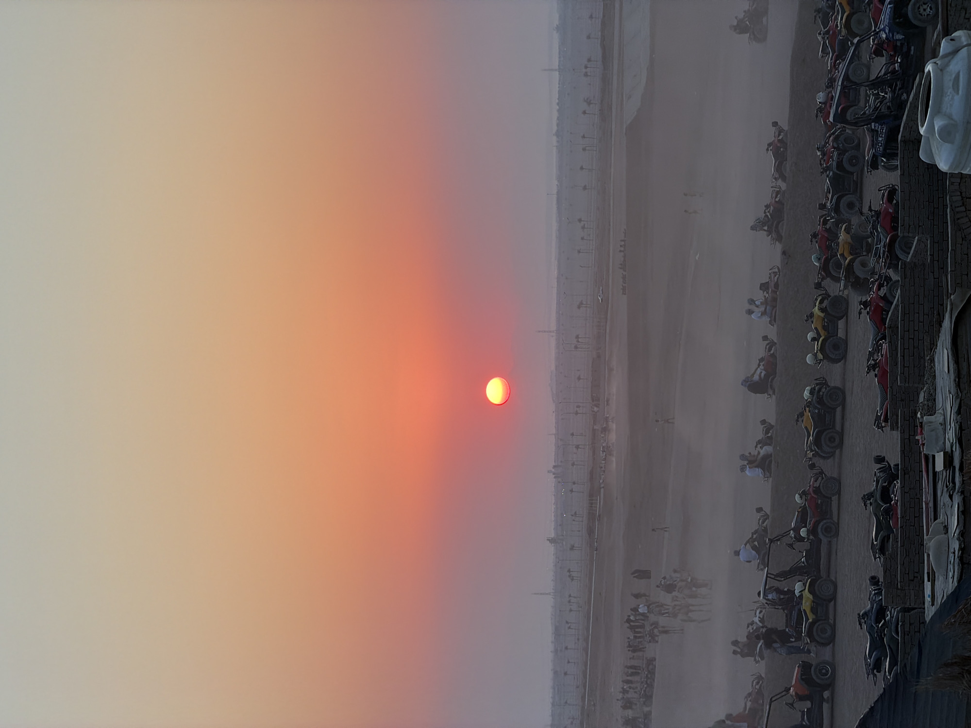This is a vertical, medium-shot photograph taken at sunset in the desert outskirts of Sharm El Sheikh, Egypt. The sun, a vibrant orange orb, is low in the sky, casting a warm, gradient glow of orange, pink, and pale yellow across the horizon. In the mid-ground, a vast, dusty desert landscape is dotted with numerous parked quad bikes and some ATVs. Several groups of people are visible, some gathered near the vehicles, while others appear to be riding or preparing to ride. In the further distance, a line of palm trees and some industrial structures are faintly visible against the hazy sky, suggesting the proximity of the city. The foreground features a closer view of more parked quad bikes, some with people seated on them. The overall atmosphere is one of leisure and adventure, with the desert and sunset providing a dramatic backdrop.