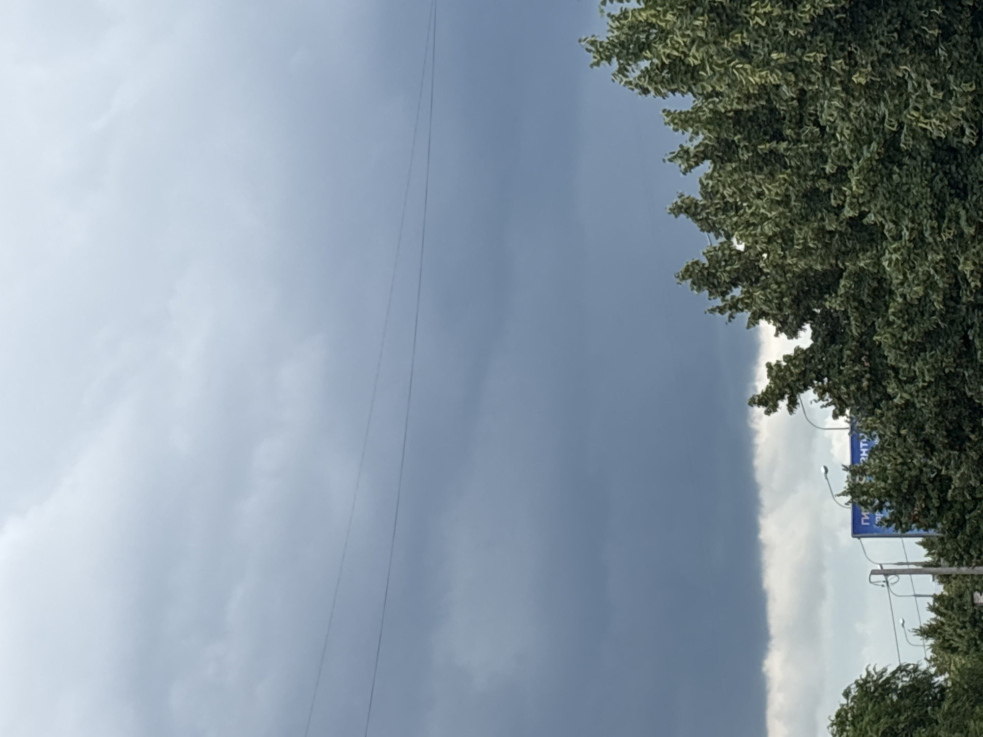 The image shows a view of the sky over Kazan, Russia, dominated by heavy, dark clouds, suggesting an impending storm. In the foreground, lush green trees frame the bottom portion of the image. Partially visible behind the trees are streetlights and a blue billboard with Cyrillic text, likely an advertisement. Power lines are visible stretching across the sky. The overall scene conveys a sense of impending weather change.