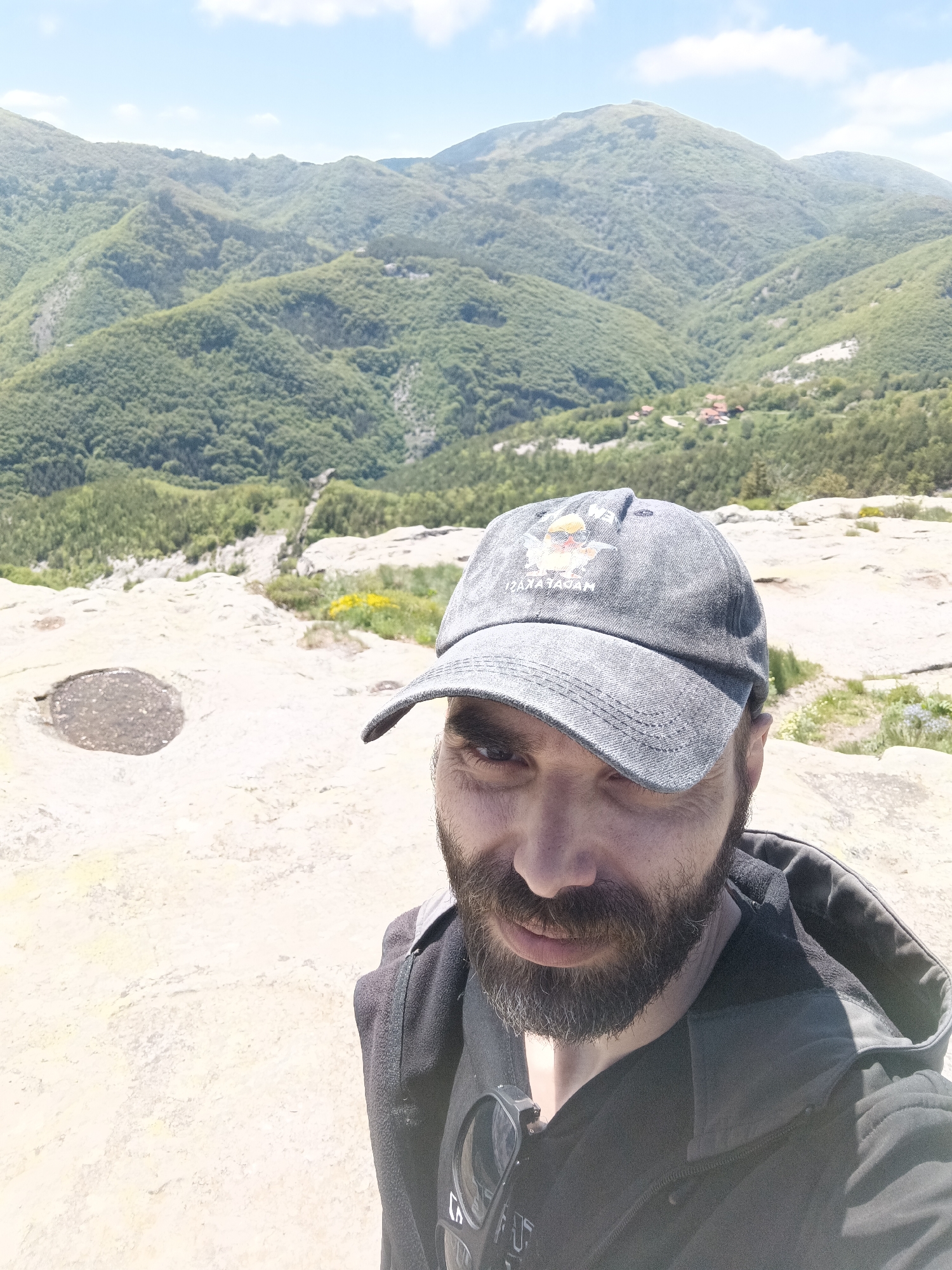 The image is a selfie taken by a man with a beard and wearing a grey cap. He is standing on a rocky outcrop with a mountain range in the background covered in lush green vegetation. The location is Sini Vrah, Bulgaria. The man is wearing a dark-colored hoodie and a black t-shirt underneath. The rocky terrain has a circular depression filled with water. The sky is light blue with some clouds.