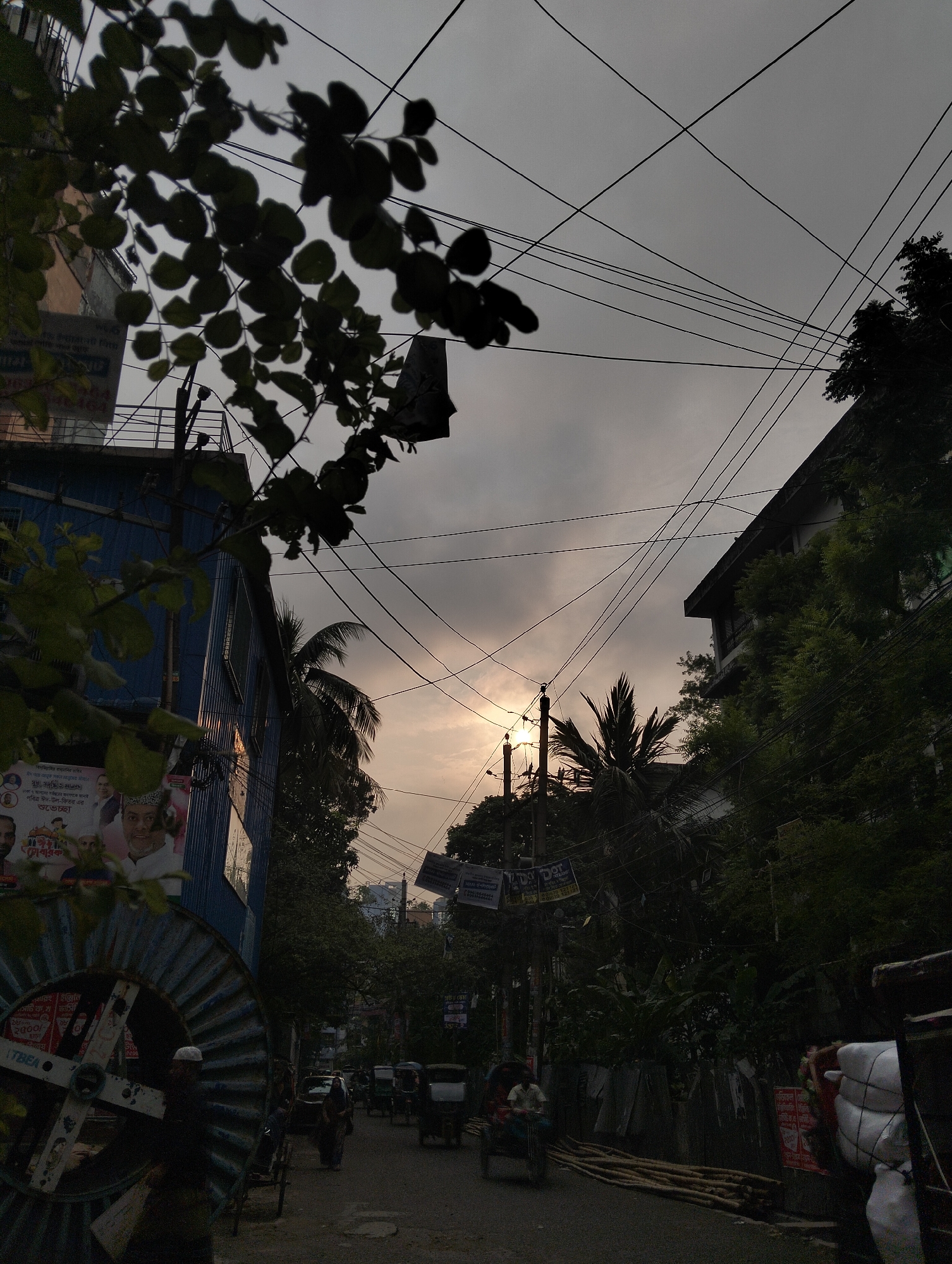 The image portrays a street scene in Dhaka, Bangladesh, under a cloudy sky with the sun faintly visible. Power lines crisscross the sky between buildings, partially obscured by silhouetted leaves and branches in the foreground. On the left, there is a blue building with posters and what appears to be a large, discarded cable reel. The street is lined with trees and various shops, with a few rickshaws and other vehicles moving along it. Several people, including rickshaw drivers and pedestrians, are visible on the street. There's a general sense of urban activity.