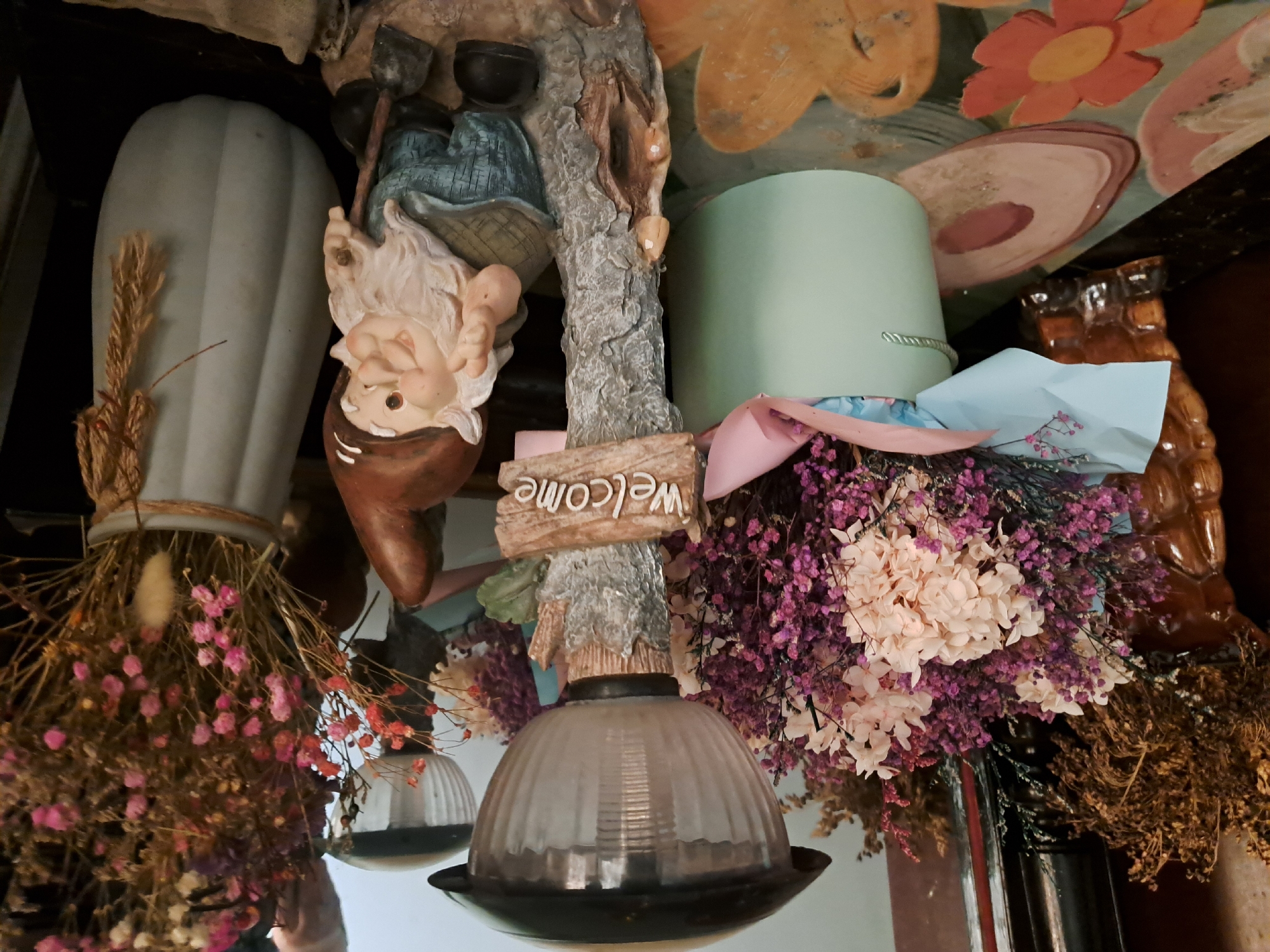 The image shows a garden gnome figurine standing next to a solar-powered lamp. The gnome is wearing a pointed hat, a checkered shirt, and blue pants, and is holding a small shovel. A "Welcome" sign is attached to the lamp post. Behind the gnome and lamp, there are bouquets of dried flowers in various colors, including pink, purple, and beige. One bouquet is in a light green cylindrical pot, and another is in a light gray vase. The scene appears to be an outdoor decorative setting, possibly a garden or a porch. The background suggests an indoor environment with dark furniture and a reflection in what appears to be a mirror or glass surface. The specific location in Khujand, Tajikistan, is not determinable from the visual information provided in the image.