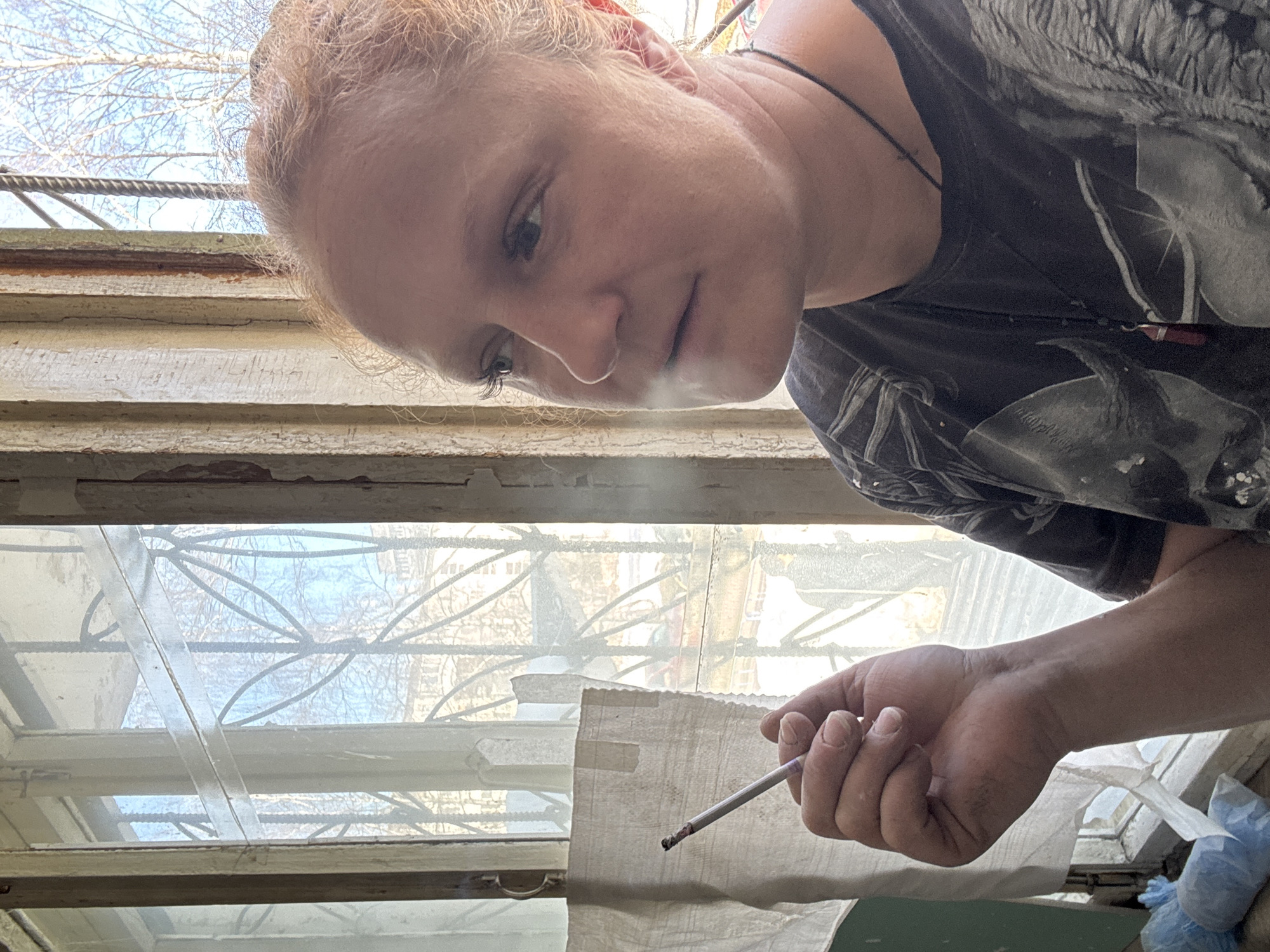 The image depicts an individual, presenting as female, indoors next to a window in Chaikovsky, Russia. The individual is shown in a profile view, facing right with a slight upward gaze. They possess light reddish-blonde hair styled away from the face, and light-colored eyes. A dark t-shirt with a grey and white print, along with a dark cord necklace featuring a reddish pendant, are worn. The left hand, positioned centrally, holds a lit cigarette between the thumb and forefinger, emitting visible smoke.
The background features a multi-pane window with worn wooden frames showing peeling paint, behind which dark, ornate security bars are visible. A light-colored fabric strip is taped to the lower-left interior window frame. Light blue plastic bags are present on a ledge beneath the window. Outside, bare deciduous trees and distant urban buildings are visible against a clear blue sky, indicating natural light conditions consistent with a cold season.