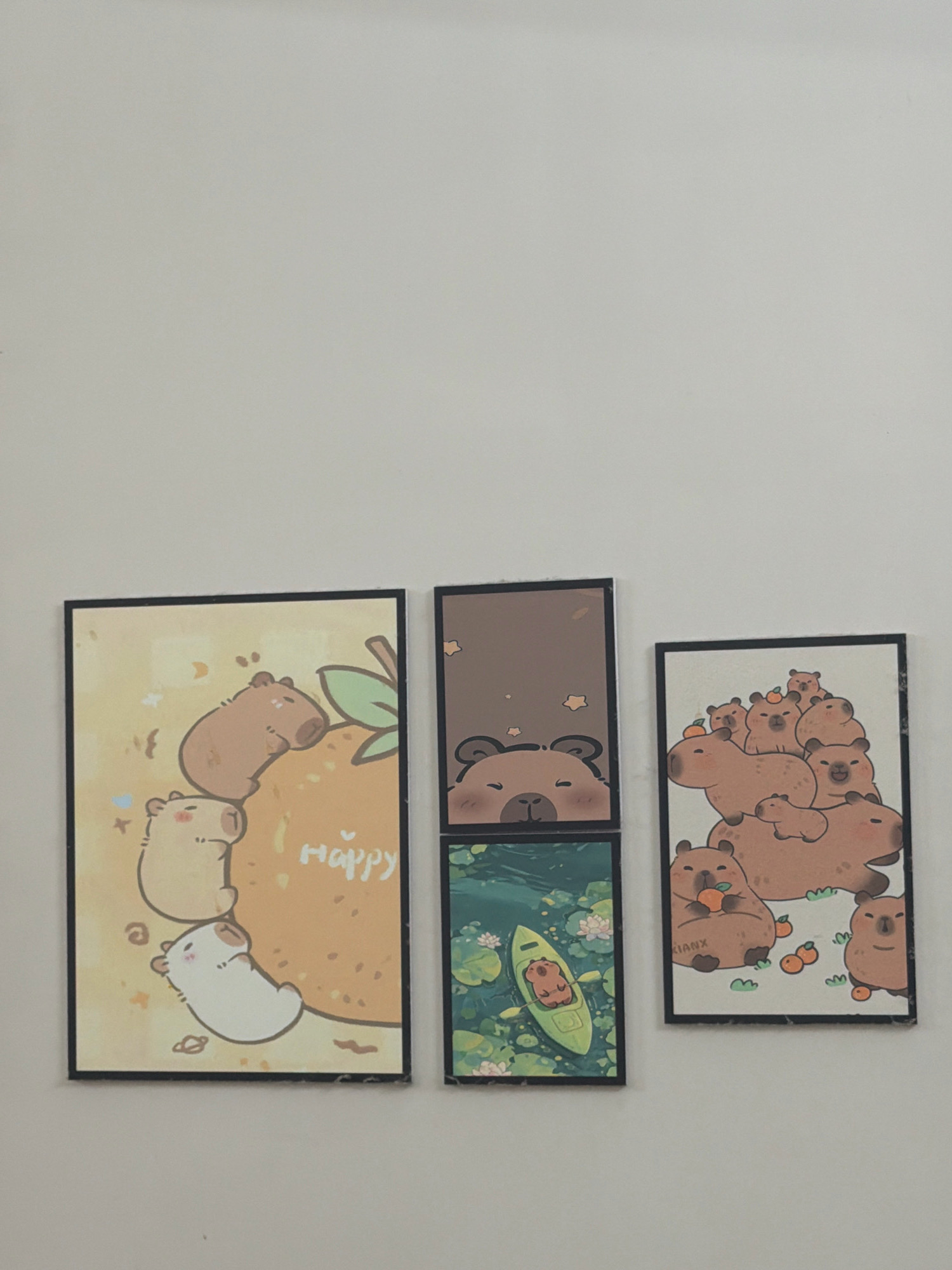 The image shows three framed prints hanging on a plain, light-colored wall. The prints are arranged in a row with a vertical gap between the leftmost and the other two, and a horizontal gap between the middle and rightmost prints.
The leftmost print is a vertical rectangular frame with a pale yellow background. It features several cartoon capybaras. The top capybara is brown and appears to be resting on an orange-brown mound with a green leaf above it. Below it, another capybara, lighter in color, is positioned facing left. The word "Happy" is written in white script on the orange-brown mound.
The middle print is a vertical rectangular frame divided into two square sections. The top section has a brown background with small yellow star-like shapes and depicts the head and shoulders of a cartoon bear peeking over the bottom edge. The bottom section is a vertical rectangular frame with a green background, depicting a body of water with lily pads and flowers. A small, yellow kayak with a capybara inside is floating on the water.
The rightmost print is a vertical rectangular frame with a white background. It displays a cluster of cartoon capybaras, varying in size, piled on top of each other. Some of the capybaras have small orange fruits near them. The word "KIANX" is written in small black letters on the bottom left capybara.
The image is taken from a slightly angled, eye-level perspective, looking up at the wall. There are no visible people or specific identifiable landmarks that would suggest a location in Nha Trang, Vietnam. The environment appears to be an indoor setting with a blank wall.