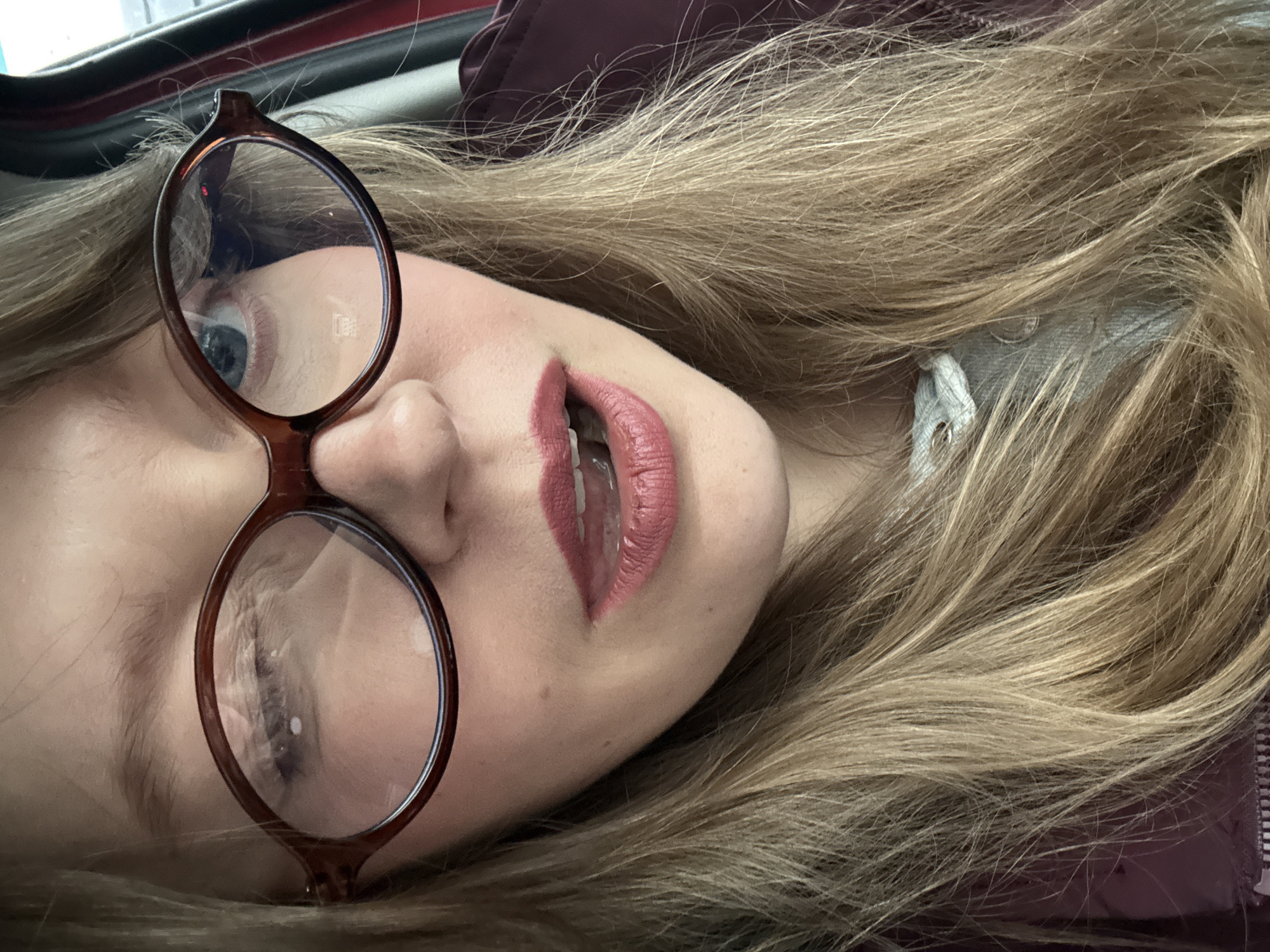 A fair-skinned person with blond hair and wearing brown-framed eyeglasses is lying down. The person has pale pink lipstick. The environment includes a red surface and a light gray drawstring. The location is not identifiable as Izhevsk, Russia.