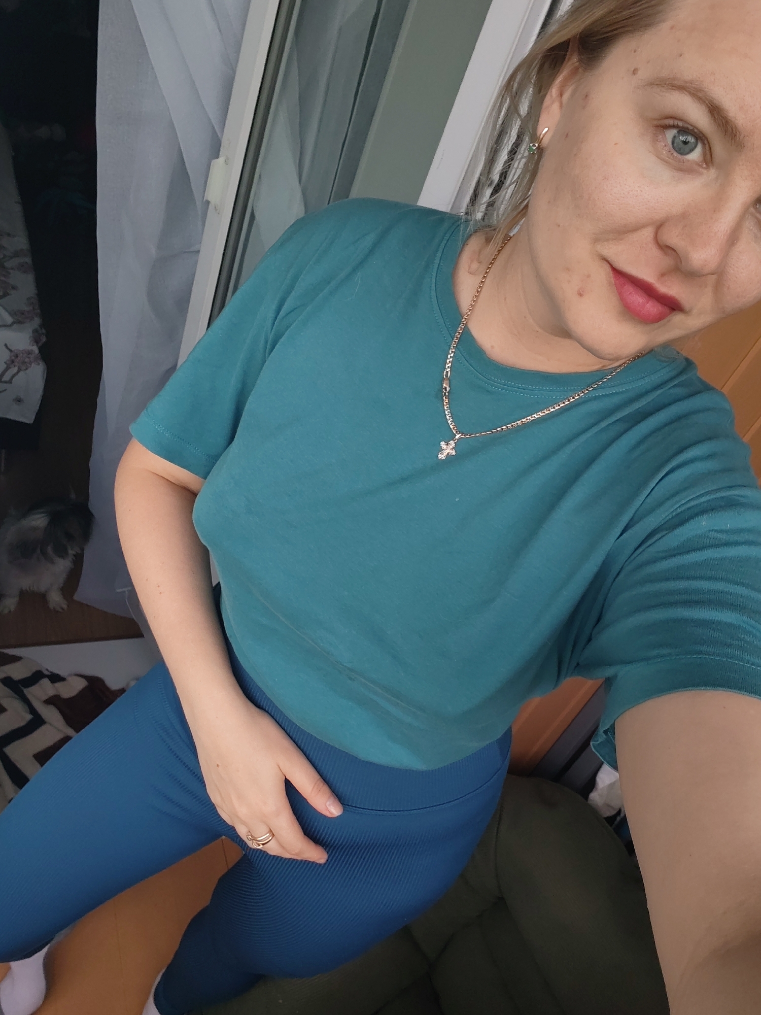 This media file is a portrait-oriented selfie of a woman. She has blonde hair pulled back, blue eyes, and is wearing a teal t-shirt and blue leggings. A gold necklace with a cross pendant is visible around her neck. She is wearing a ring on her left hand, which is resting on her leg. The background is partially visible and includes a window with white curtains, a wooden floor, and what appears to be a dog in the distance. The lighting suggests it is daytime. The woman's expression is neutral. There is no visible text in the image. The location context provided is Izhevsk, Russia.