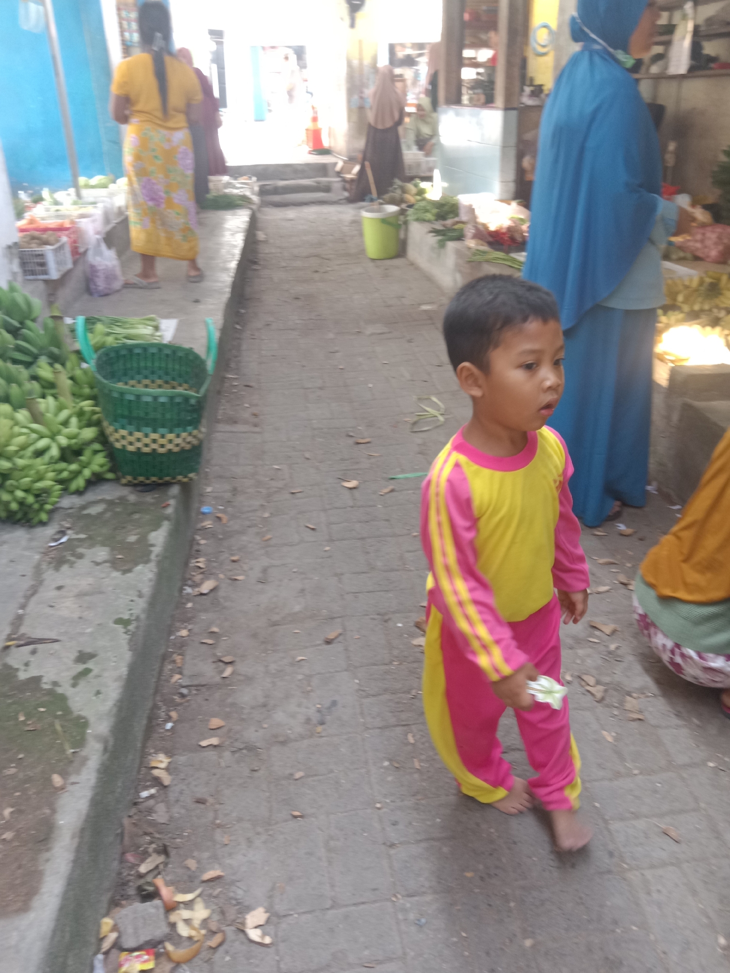 The image captures a street scene in Klepek, Indonesia, likely a market area. In the foreground, a young child with dark hair is walking barefoot, wearing a bright yellow long-sleeved shirt with pink stripes and matching pink pants with yellow accents. The child is holding a small white object in their right hand. To the left, a display of green bananas is visible on a raised concrete ledge, with a green and yellow woven basket next to them. Behind the child, several adults are present. One person, partially visible, wears a blue hijab and a blue tunic. Further back, another individual is seen from behind, wearing a yellow top and a patterned skirt, with their hair in a ponytail. A person in a light brown hijab is also present. The ground is paved with grey stone tiles, and scattered leaves and debris are visible. The background shows more market stalls with goods and structures typical of an Indonesian street.
