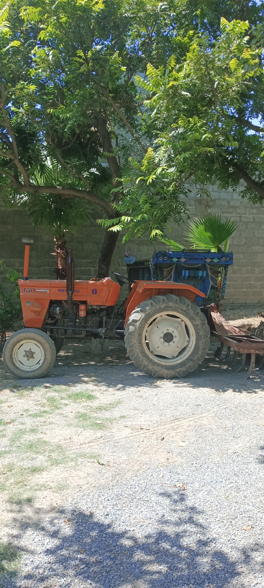 An orange New Holland 480 Special tractor is parked on a gravel surface in Katlang Tehsil, Pakistan. A large tree with dense green foliage partially shades the tractor. A brick wall is visible in the background, with a palm-like plant near the wall. The tractor has a blue decorated frame around the operator's area, and a harrow implement is attached to the rear.