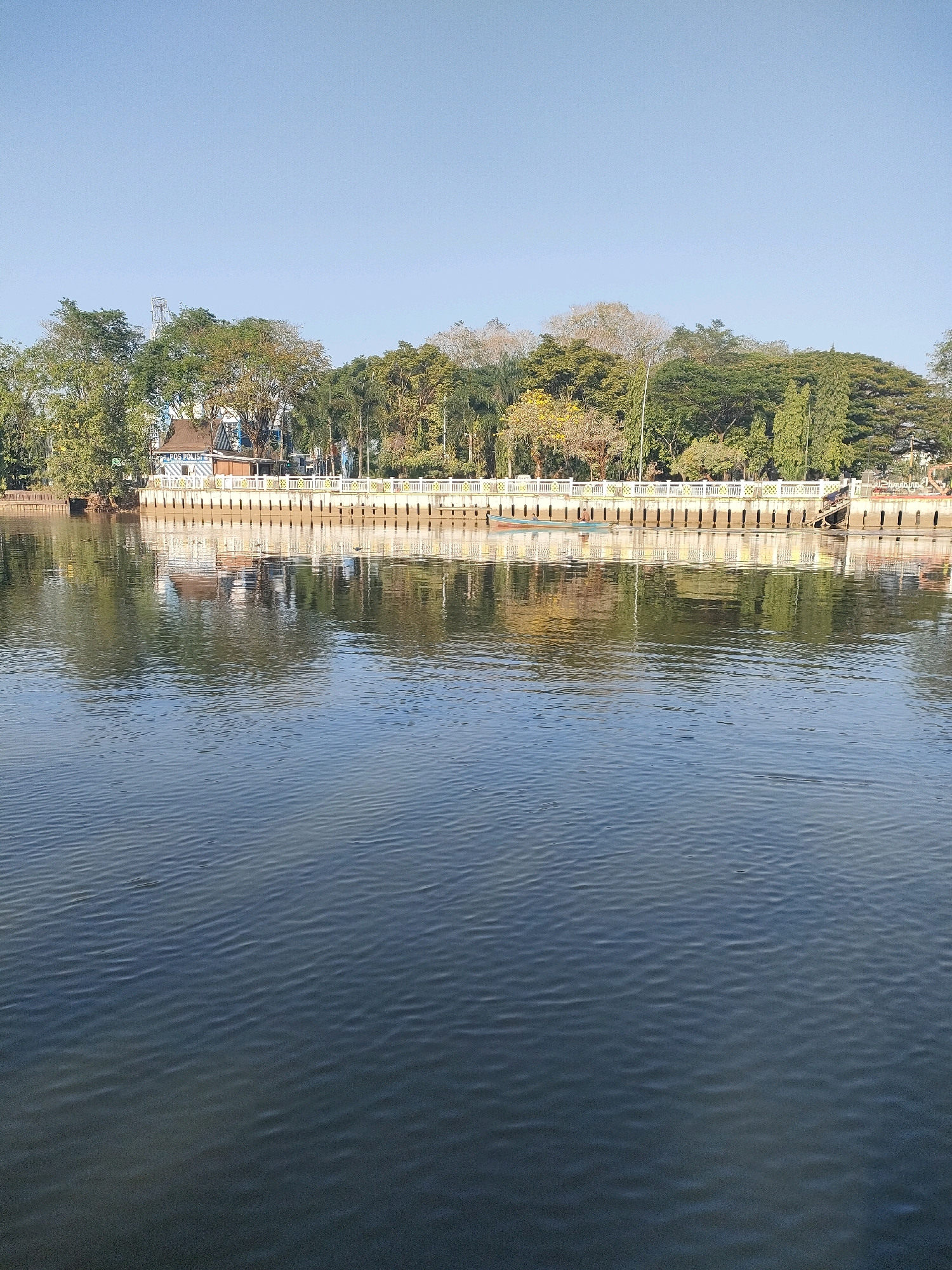 This is a daytime, eye-level, full-shot photograph of the Barito River in Banjarmasin, Indonesia. The river occupies the foreground, with gentle ripples disturbing its dark, reflective surface. In the mid-ground, a long, ornate white embankment separates the river from a line of trees and a building. This embankment features a decorative railing with yellow accents and a series of evenly spaced pillars. Behind the embankment, a variety of trees with green foliage can be seen, some with yellow flowers. A multi-story building with a blue roof and brown walls, identified by the text "POS POLISI" (Police Post) on its side, is visible nestled amongst the trees. There are a few people in the scene. One person is in a small, dark-colored boat with a light-colored stripe, drifting near the embankment. Another person, wearing a blue shirt and possibly a hat, is standing in the boat. Further to the right, several figures are visible walking along the embankment. The sky above is clear and a light blue, indicating fair weather. The reflection of the embankment and trees is clearly visible in the water, adding depth to the scene.