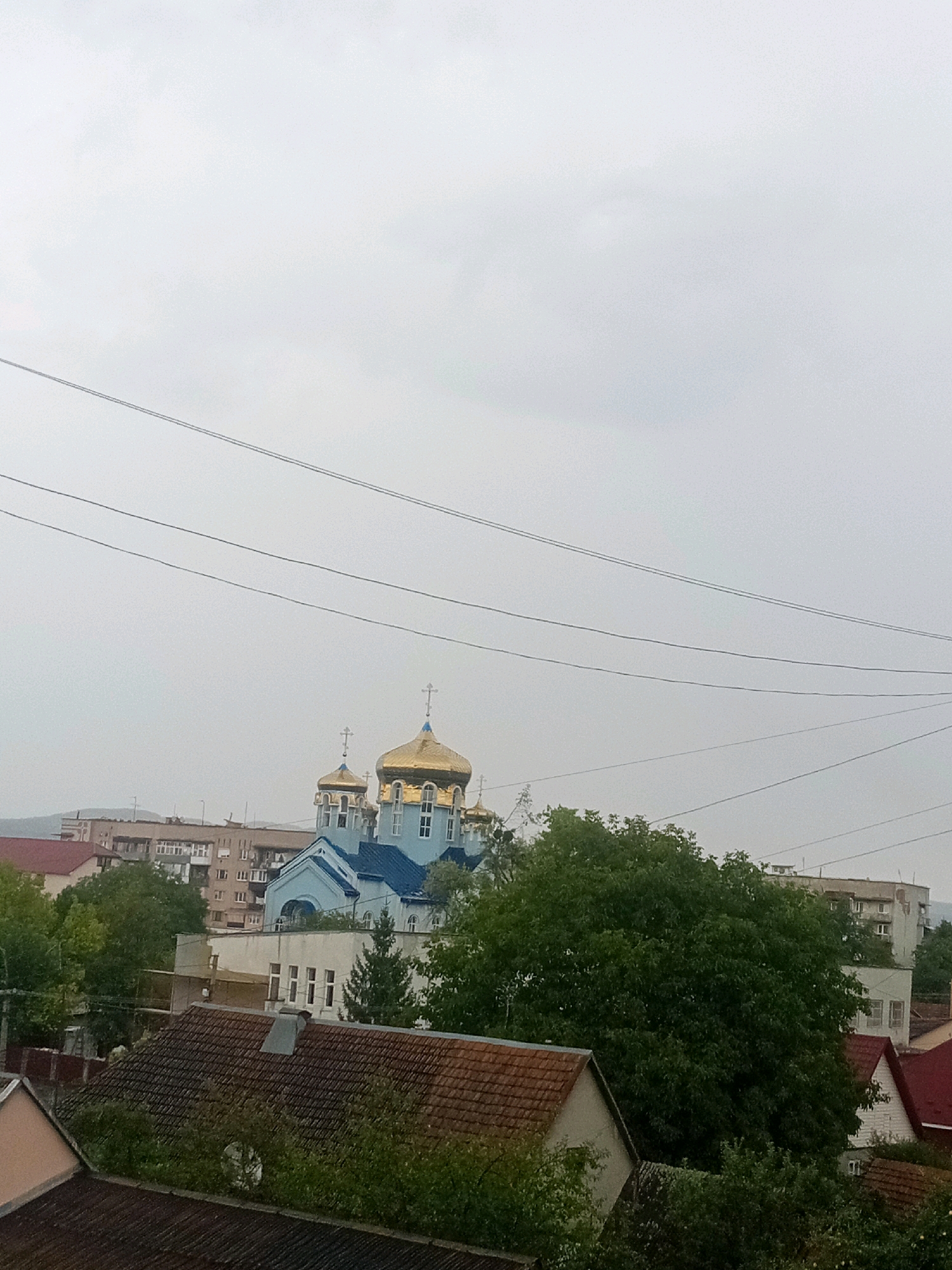 The image captures a view of Tiachiv, Ukraine, dominated by a prominent blue church with golden domes. The church is nestled amongst trees and other buildings, with a gray, overcast sky overhead. Several power lines crisscross the upper portion of the frame. In the foreground, there are rooftops of residential buildings, some with red tile and others with darker shingles. The middle ground shows the church and lush green trees, while the background consists of apartment buildings and a distant hillside. The scene appears to be a residential area with a notable religious landmark.