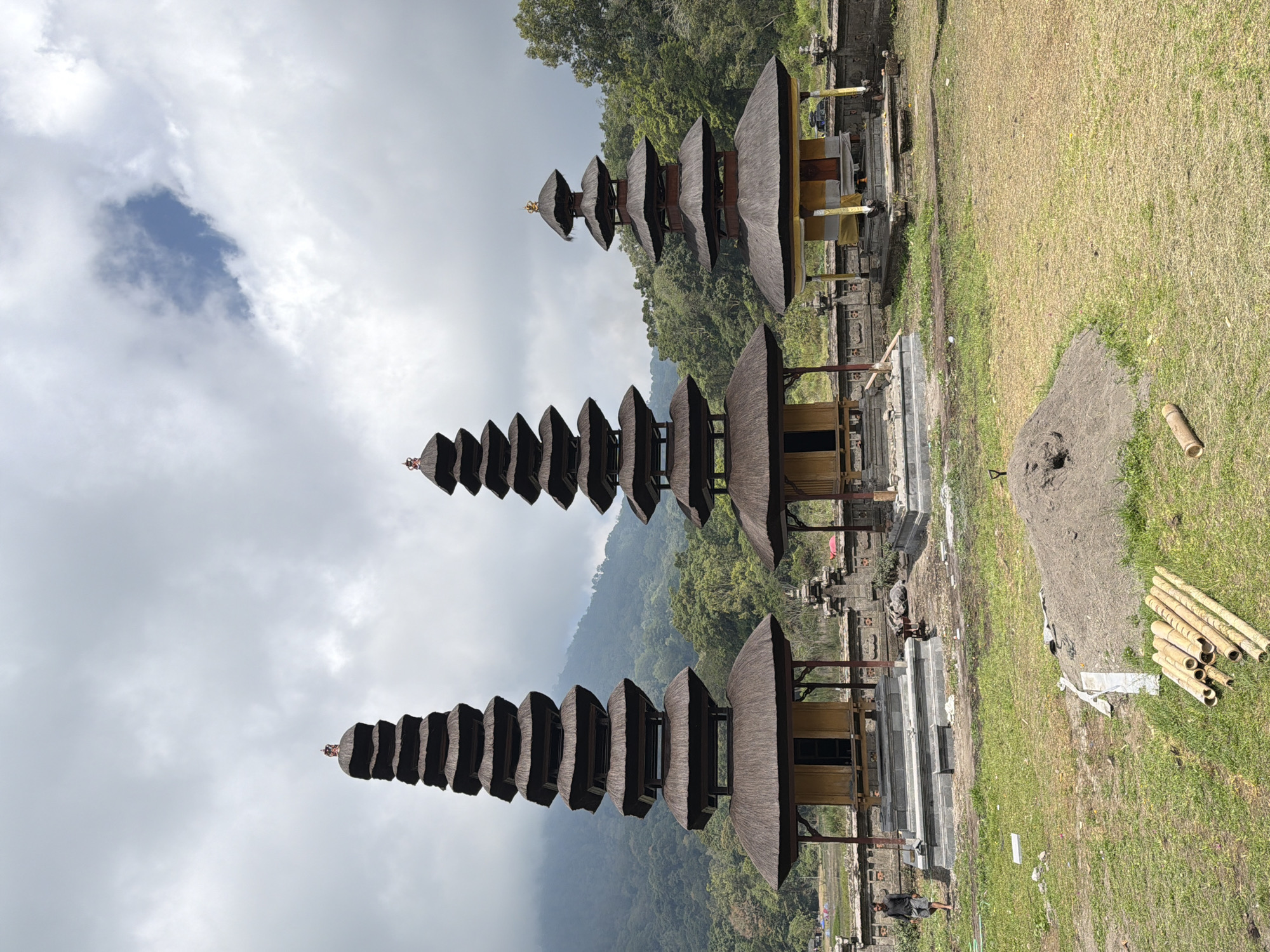 In Banjar, Indonesia, a serene scene unfolds at a temple complex with three distinctive meru towers. The towers, each with multiple tiered roofs thatched with straw, stand tall against a backdrop of lush green hills and a sky filled with dramatic clouds. The central tower is the tallest, followed by the one on the left, and then the one on the right. In the foreground, a man in a dark shirt and shorts walks on the grassy ground. Scattered bamboo poles lie on the grass, hinting at ongoing construction or maintenance. A pile of sand is also visible, further suggesting activity. The temple grounds are enclosed by stone walls, and the architecture of the meru towers, with their layered roofs and open-sided structures, is characteristic of Balinese Hindu temples. The overall atmosphere is peaceful, with a sense of historical and spiritual significance.