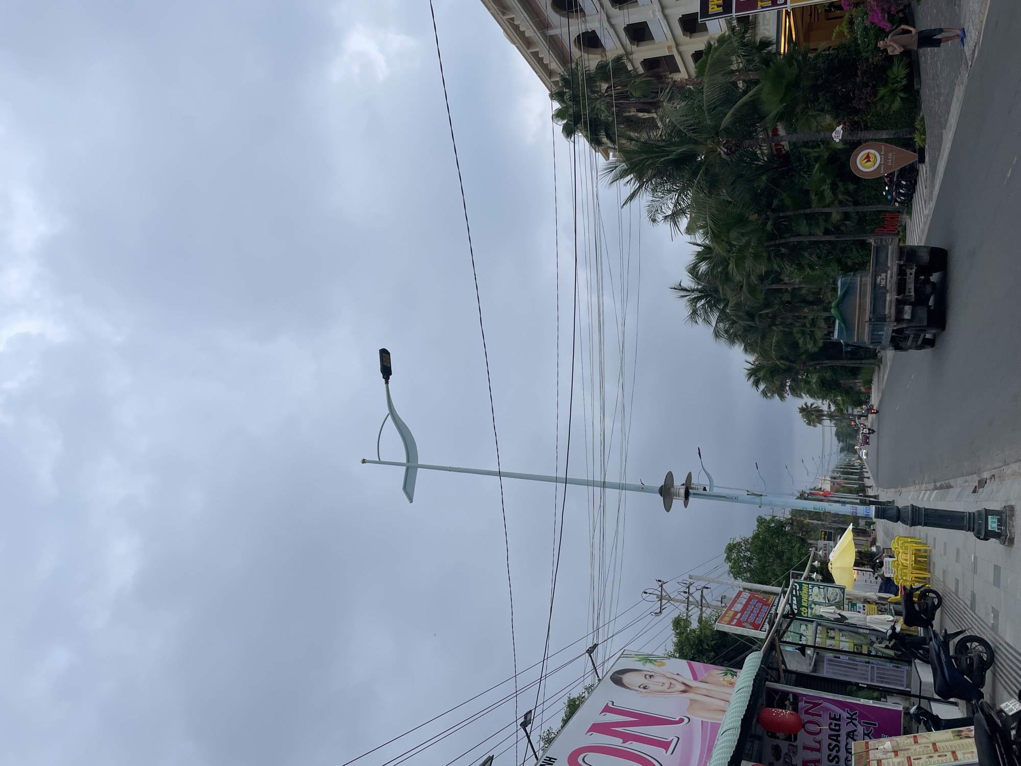 The image displays a street scene in Phu Thuy Ward, Vietnam, on an overcast day. The perspective looks down a long road, with sidewalks flanking both sides.
On the left side of the road (foreground), a series of commercial establishments line the sidewalk. A large pink sign prominently features the word "SALON," with a woman's face depicted on it. Partially visible text beneath suggests services like "MASSAGE" and "TAXI." A yellow umbrella shades an area with yellow plastic chairs, indicating an outdoor eatery or cafe. Several motorbikes are parked along the sidewalk. A tall, modern street light pole with a distinct curved design stands in the foreground, with numerous utility lines strung overhead, crisscrossing the sky. Further down the street, a more traditional black street light pole is visible.
On the right side of the road, a multi-story building with ornate balconies and brown shutters rises above a dense line of tall palm trees. A large, dark truck is seen on the road, driving away from the viewer. A wooden, pointer-shaped sign, featuring a circular logo and partial text like "Resort & Spa," "BIEN VUONG," and "BEACH," is placed among the palm trees. In the distance, a person is visible walking on the sidewalk.
The sky is uniformly cloudy and grey, suggesting an overcast day. The scene captures everyday urban life, characterized by a mix of businesses, tropical greenery, and bustling street activity.