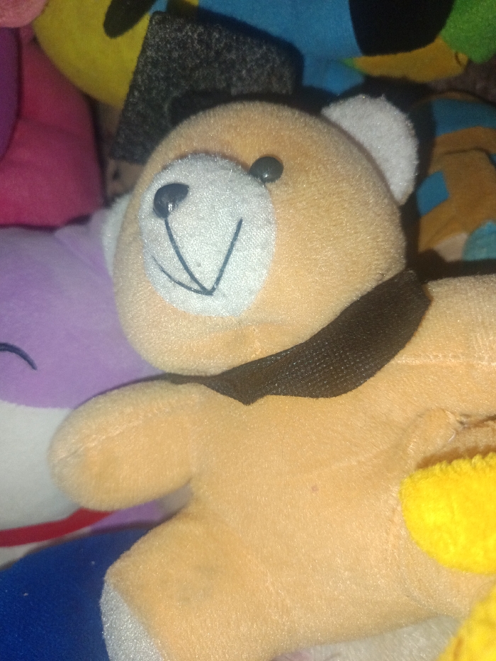 The image depicts a close-up, angled view of multiple plush toys densely arranged. Centrally featured is a light tan plush bear with a white snout, black button eyes, a stitched black nose, and a stitched V-shaped mouth. White fabric forms its inner ears, and a black fabric ribbon is tied around its neck. This bear is situated amongst and partially obscures other plush toys. Visible surrounding elements include a purple plush with a stitched line to the left of the bear, a yellow plush partially visible above the bear's head, and indistinct segments of pink, dark grey, and blue plush materials in the background. No contextual information is present within the image to determine a specific geographical location, including any details pertaining to the city of Gotha, Germany.