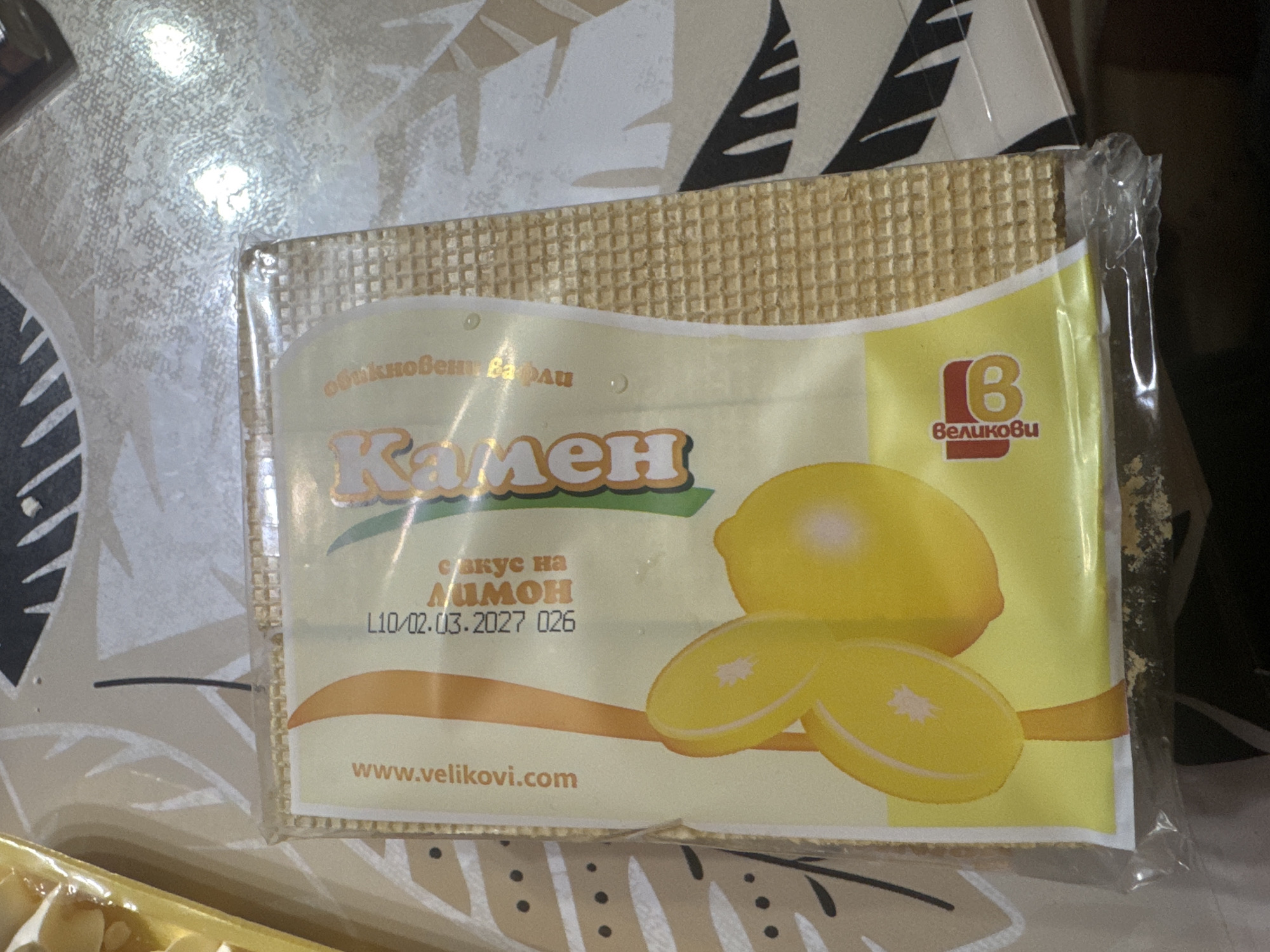 A close-up shot of a package of Velikovi "Kamen" lemon-flavored wafers. The packaging is predominantly yellow and white, with an image of a lemon and lemon slices on the front. Text on the package includes the brand name "Velikovi" and a website address, www.velikovi.com. An expiration date is partially visible: L10/02.03.2027. The wafers themselves are visible through the clear plastic packaging. The product is resting on a surface with a patterned design. The wafers are made in Sarnevo, Bulgaria.