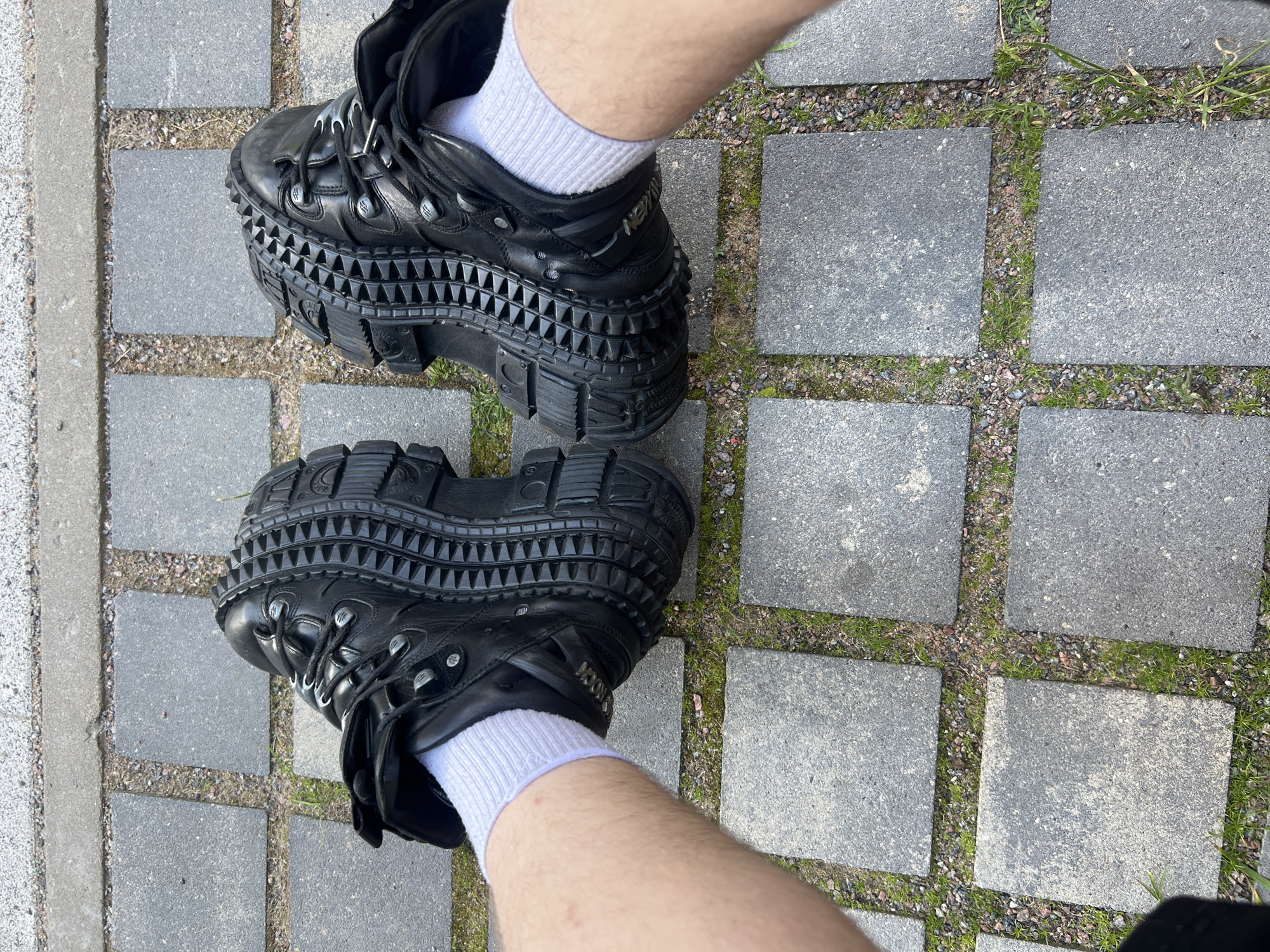 The image displays a high-angle, downward view of a person's legs and feet, clad in distinctive black platform boots and white ribbed socks, positioned on an outdoor paved surface. The boots are chunky and made of black material, likely leather, featuring an exceptionally thick, multi-layered platform sole. The side of the platform is adorned with a prominent pattern of small, uniform pyramidal studs, while the underside of the sole has a robust, lugged tread. Several metal eyelets are visible on the lacing area of the boots, and the brand name "NEW ROCK" is embossed on the lateral side of one boot. The white ankle socks provide a sharp contrast to the dark footwear. The ground beneath consists of square gray paving stones, laid with narrow gaps filled with a mixture of small gravel and patches of green grass or weeds, suggesting a walkway or garden path. The scene is illuminated by natural daylight, indicating it is daytime. The person appears to be standing, showcasing their unique footwear. The provided context situates the scene in Saint Petersburg, Russia.