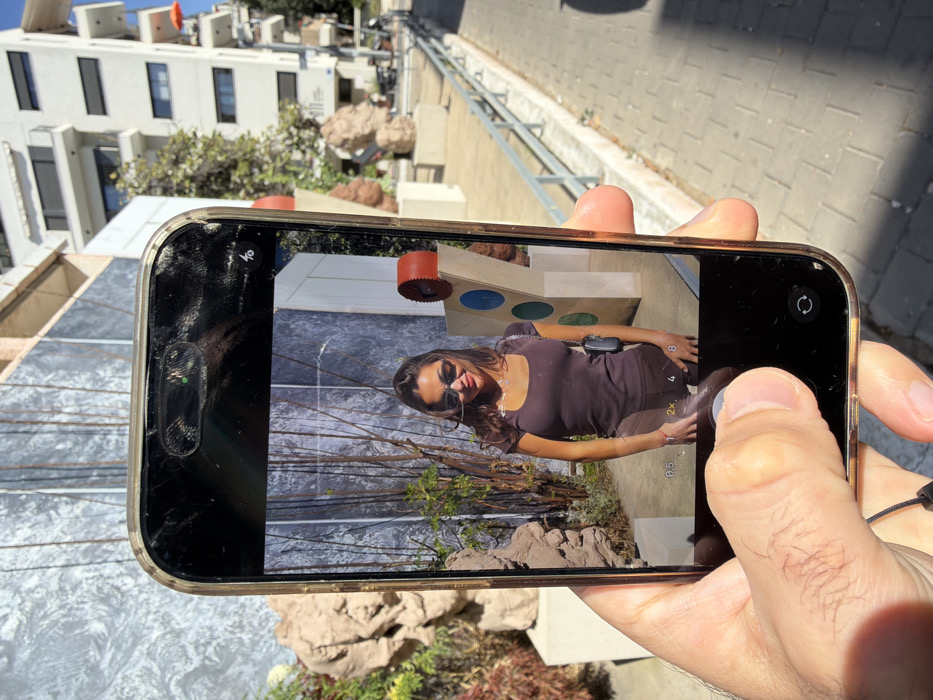 A close-up, elevated perspective shows a left hand holding a gold-framed smartphone in portrait orientation, displaying an active camera application. The phone screen exhibits multiple cracks, particularly visible across the top and near the dynamic island cutout, which shows a small green indicator light. A right thumb rests near the bottom center of the screen, positioned over the shutter button. The camera app's user interface is visible, including zoom options (0.5x, 1x, 2x, 4x, 8x) and a camera switch icon. A black lanyard or strap hangs from the phone's bottom left.
The smartphone's screen displays a young woman standing outdoors, smiling towards the camera. She has dark hair, wears dark sunglasses, a dark brown short-sleeved top, dark bottoms, and a small dark shoulder bag. She has a necklace and a bracelet visible. Behind her on the screen is a textured grey wall or vertical surface, appearing like rough concrete or stone, alongside green foliage and various art installations.
The real-world background behind the phone largely mirrors the scene depicted on the screen. To the left, a large, abstract, reddish-brown sculpture with organic shapes is partially visible, consistent with contemporary public art. The textured grey wall extends upwards. To the right, a paved pedestrian pathway extends into the distance, bordered by a low metal railing. Beyond the pathway are several multi-story white residential buildings with dark-framed windows and balconies, under a clear blue sky. One building features a distinctive orange umbrella on a balcony. The distinct art installations and the architectural style of the buildings indicate the location as the outdoor plaza adjacent to the Suzanne Dellal Center for Dance and Theater in the Neve Tzedek neighborhood of Tel-Aviv, Israel. The scene is brightly lit by sunlight, casting visible shadows.