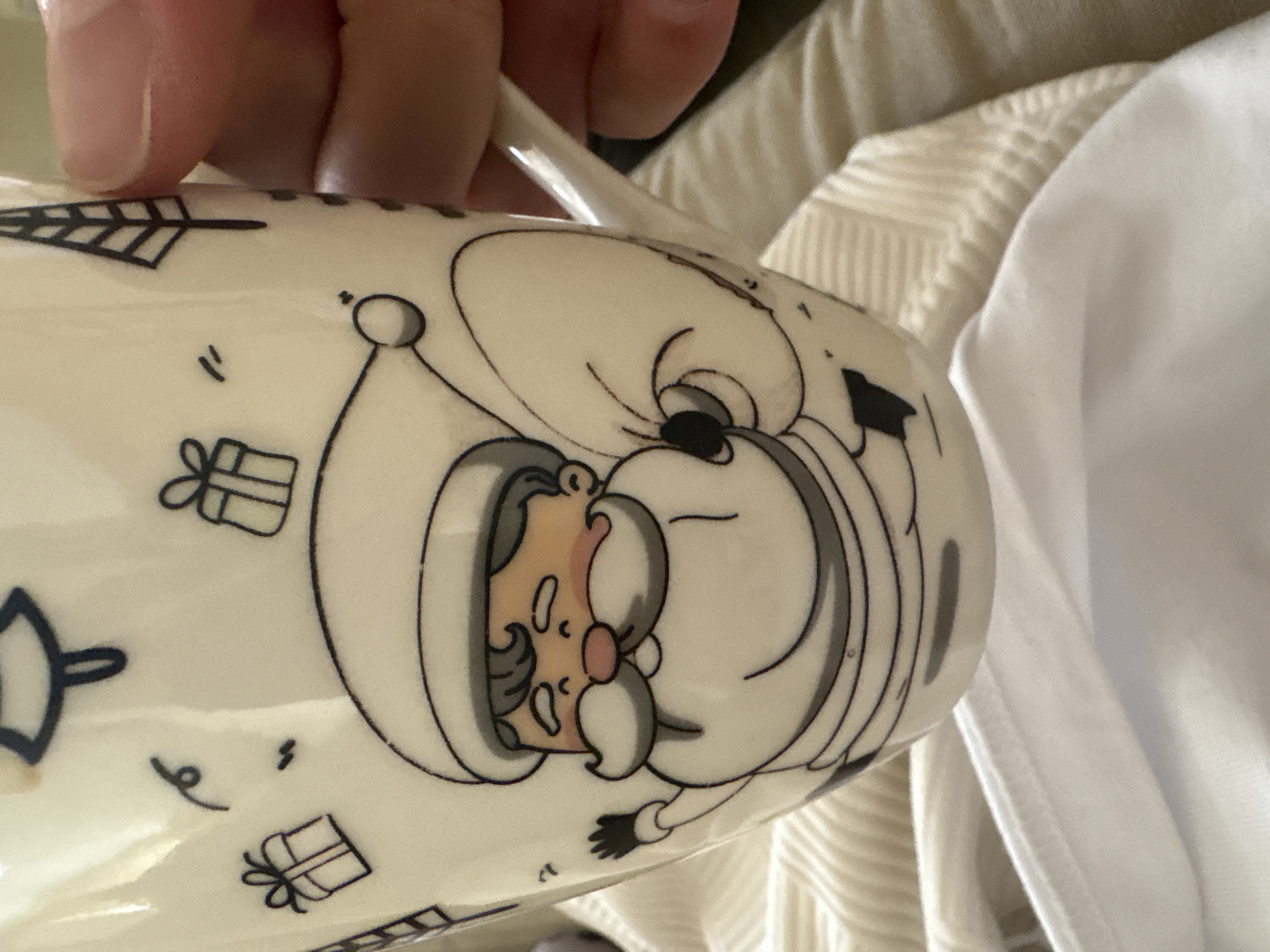 A partial view of a white mug featuring a cartoon depiction of Santa Claus is displayed. The mug is adorned with additional designs, including stylized Christmas trees and gift boxes. An individual's hand is visible, holding the mug by its handle. The background shows light-colored, folded textiles. There is no indication of the mug being in Mikrevo, Bulgaria.