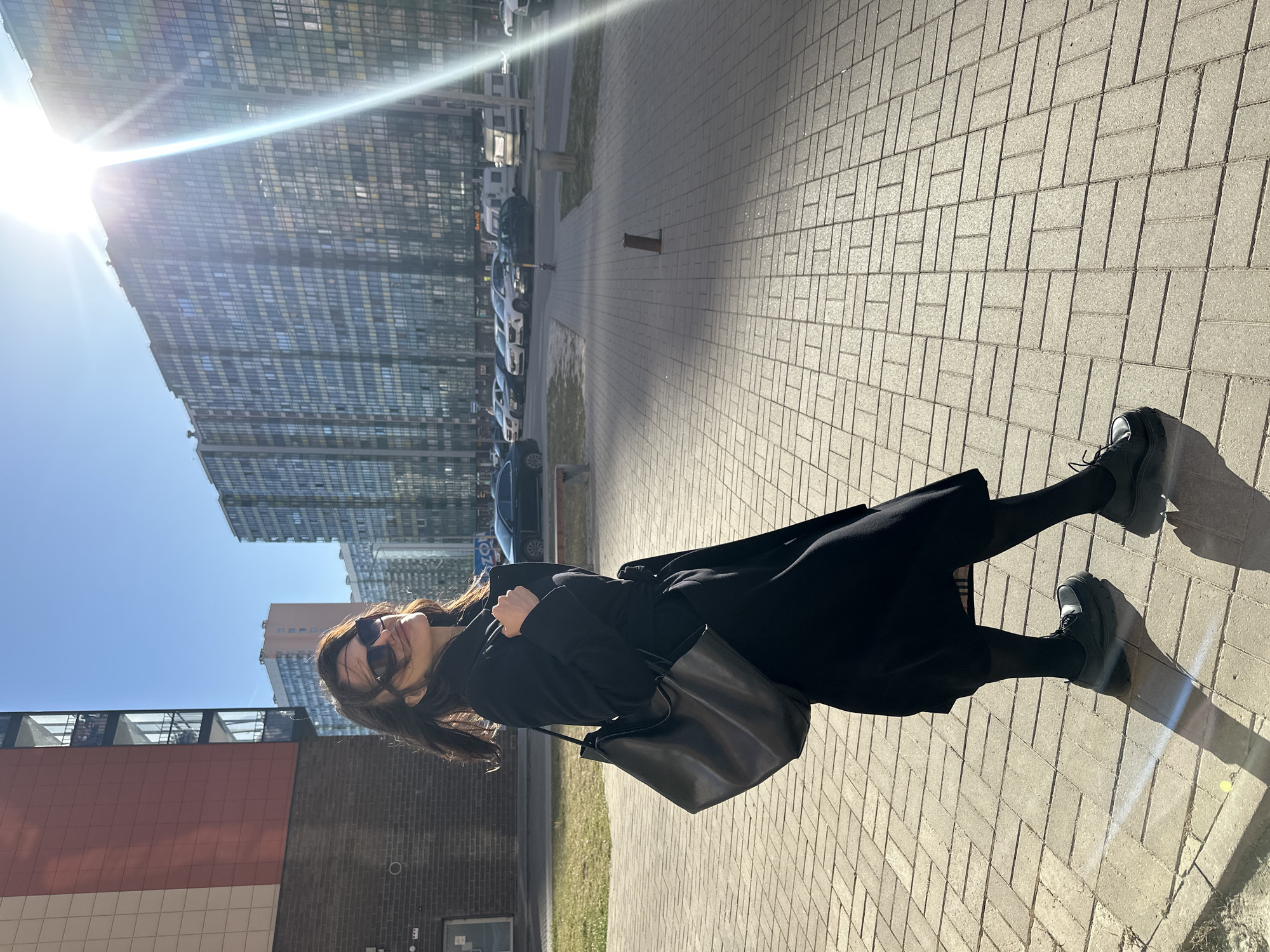 The image captures a woman standing on a paved sidewalk in a modern urban setting under bright sunlight. The woman, positioned slightly to the right of the frame, has dark hair and wears sunglasses, a long black coat or dress, black tights, and black platform shoes. She carries a black, seemingly leather, shoulder bag and is looking towards the viewer with a slight, relaxed smile.
Behind her, a street with several parked cars is visible. The background is dominated by contemporary architecture, including a prominent tall building with numerous reflective glass windows that brilliantly reflect the sunlight, creating a strong sun flare in the upper left portion of the image. Other buildings with red and brown facades are also present. The sky is clear and blue. The strong sunlight casts distinct shadows across the light-colored, rectangular block paving of the sidewalk.
A blue sign with white text, appearing to read "OZON," is faintly discernible on one of the buildings in the middle distance. The scene conveys a bright, casual daytime moment in a city environment, indicative of a modern district like Murino, Russia.