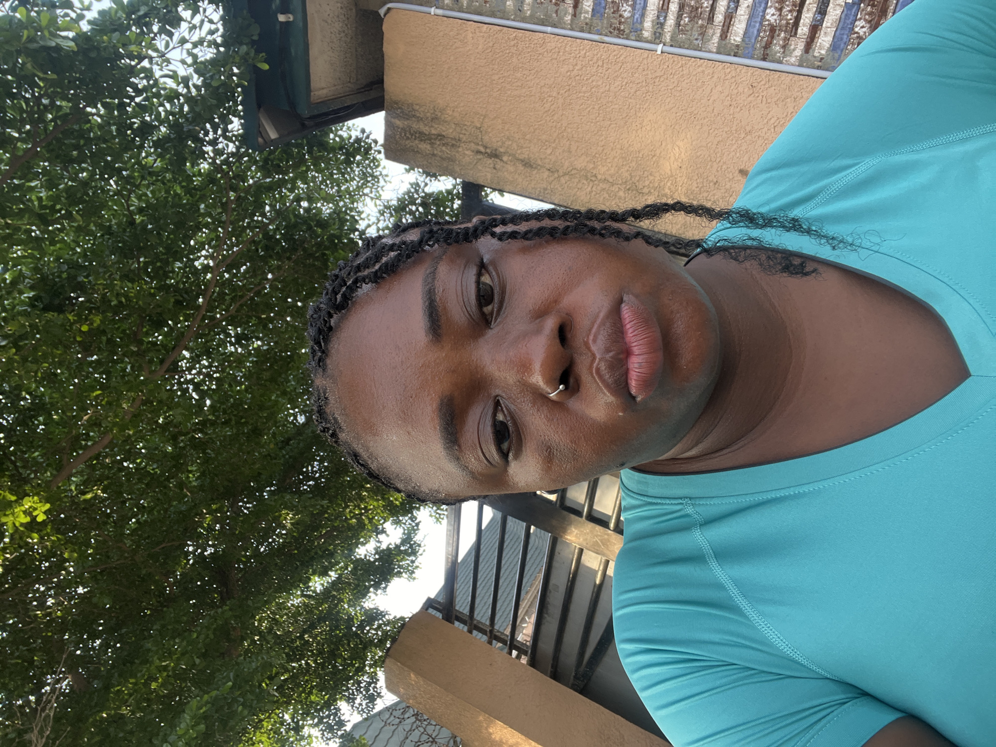 A medium shot captures a Black woman with dark braided hair and a nose ring in an outdoor setting. She is wearing a teal t-shirt and has her lips pursed, looking directly at the camera. Behind her, a textured beige wall with a metallic grate and some wires is visible, suggesting an urban environment. Above, lush green tree foliage fills the upper portion of the frame, with dappled sunlight filtering through. The overall impression is a candid portrait taken in a city like Abuja, Nigeria, possibly during daytime given the lighting.