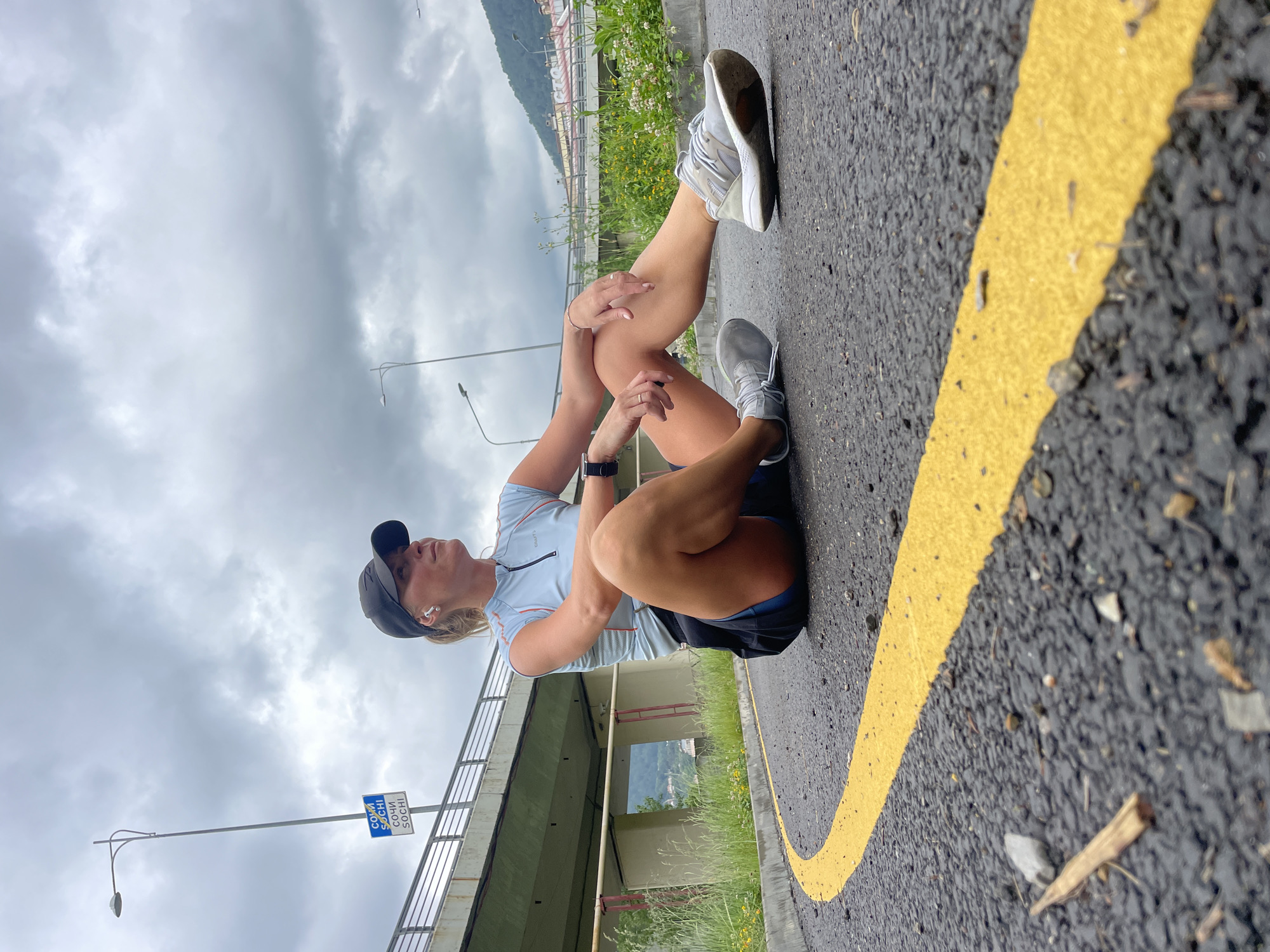 A woman is sitting on an asphalt road with a yellow line painted on it. The road is located under a bridge in Sochi, Russia, as indicated by a blue and white sign that reads "СОЧИ SOCHI" attached to a lamppost. The woman wears a black baseball cap, a light blue athletic top, dark shorts, and running shoes. She is holding her right knee with both hands. She has wireless headphones in her ears and a wristwatch on her left wrist. The environment includes a cloudy sky, vegetation growing along the side of the road, and a view of the mountains in the distance.
