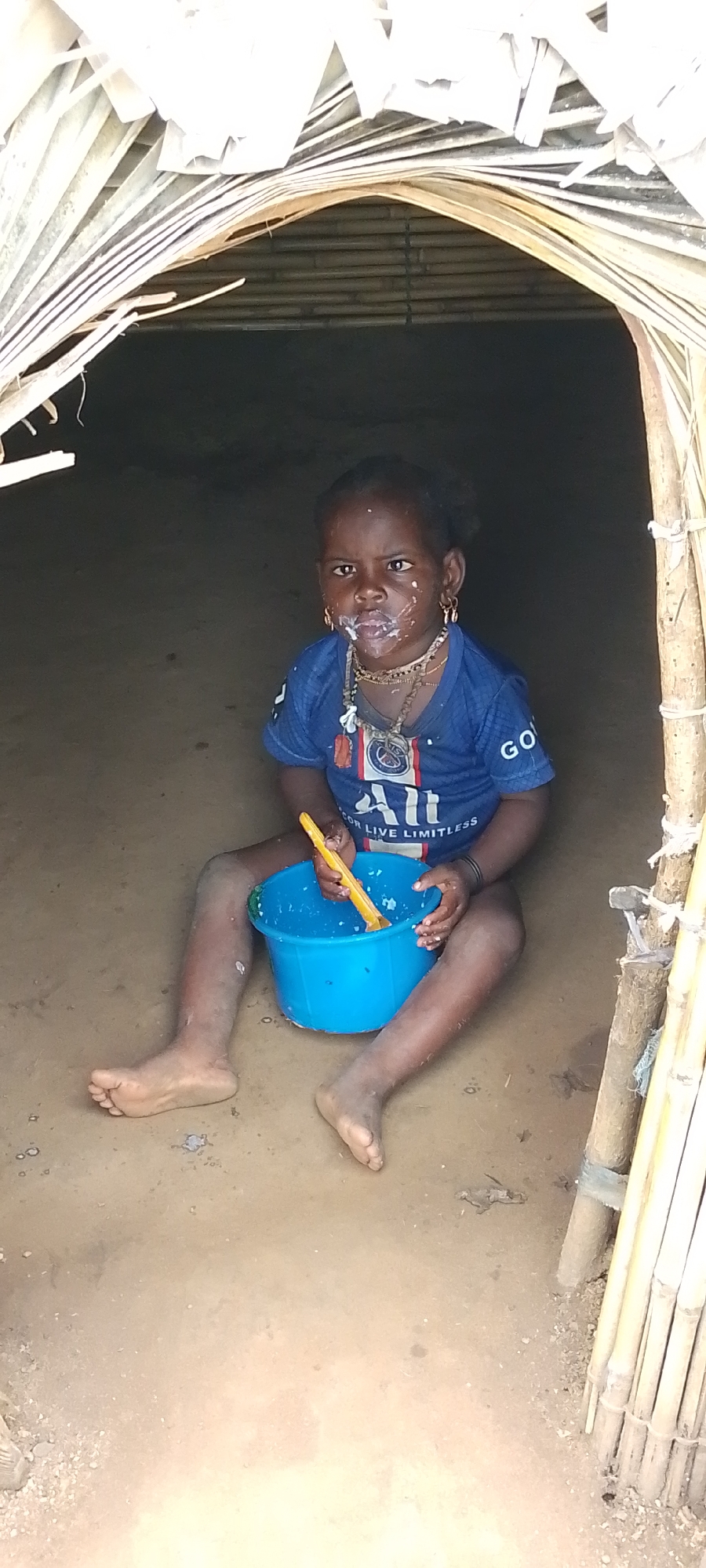 A young child sits on the dirt floor of a hut in Kamantan, Nigeria, holding a blue bowl with a yellow spoon. The child is wearing a blue t-shirt and several necklaces, and their face is smeared with a white substance. The hut appears to be constructed from natural materials, with palm leaves forming the roof and bamboo poles supporting the entrance. The lighting is dim, suggesting the photo was taken indoors.