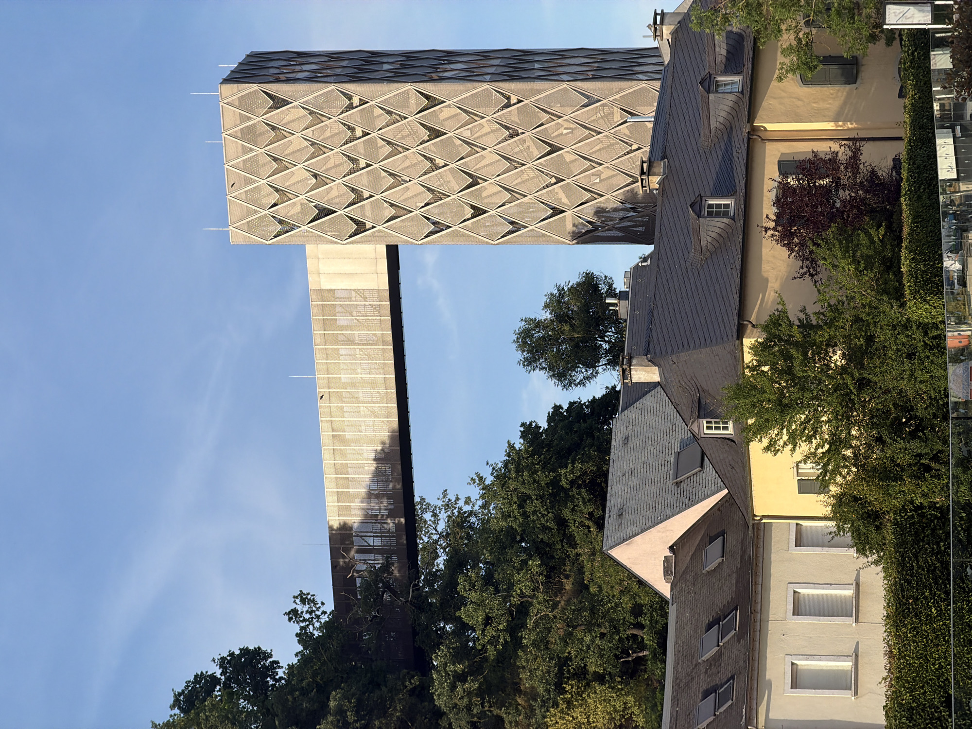 The image shows a modern observation tower with a skywalk in Fentange, Luxembourg, against a clear blue sky. The tower, which has a diamond-patterned facade, is connected by a horizontal skywalk to a structure on a tree-covered hill. In the foreground, traditional houses with dark tiled roofs are visible, some with dormer windows and chimneys. Dense green trees and hedges partially obscure the houses and the base of the tower. The overall scene depicts a juxtaposition of modern architecture with historic residential buildings and natural landscape.