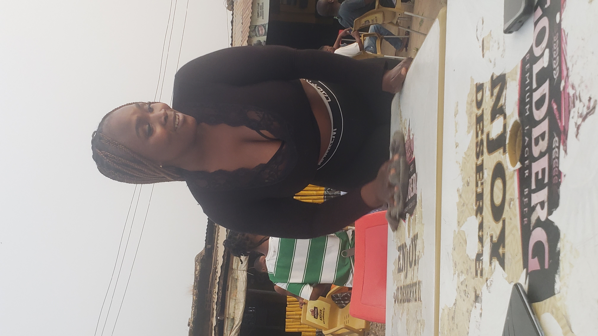 A woman with braided hair stands on the left, smiling while cleaning a table outdoors. She is wearing a black, long-sleeved, lace-trimmed top and black pants with visible waistband. A gray rag is in her right hand, wiping the white table with the word "GOLDBERG" visible on the table's surface.
Behind the woman, stacks of yellow plastic chairs are visible, along with a person in a green and white striped shirt. Other people are seated in the background, near a corrugated metal structure. The location appears to be a commercial area within Narayi, Nigeria.