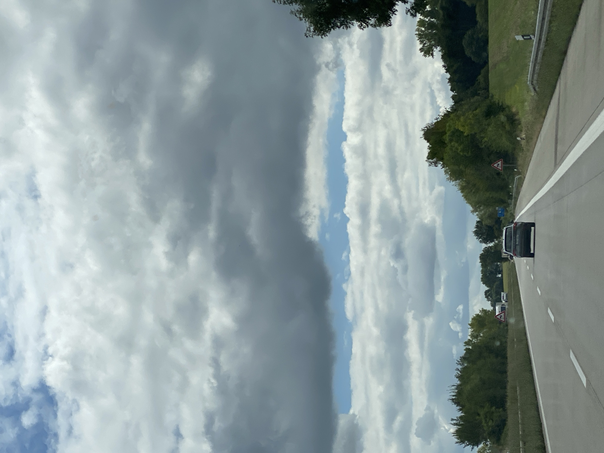 This image depicts a view from a moving vehicle on a highway, likely in or near Roßwein, Germany, based on the context provided. The sky is filled with dramatic, overcast clouds, with a narrow band of blue sky visible in the mid-ground. The highway stretches forward, with white lane markings clearly visible. A dark-colored SUV is the most prominent vehicle, driving ahead in the right lane. On the left side of the highway, there is a grassy median with a line of trees and vegetation. Several road signs are visible, including a triangular warning sign with a symbol of animals, and a sign indicating a caravan or trailer. On the right side of the highway, a grassy embankment rises, topped with more trees. The overall impression is of a typical highway scene under a cloudy sky.