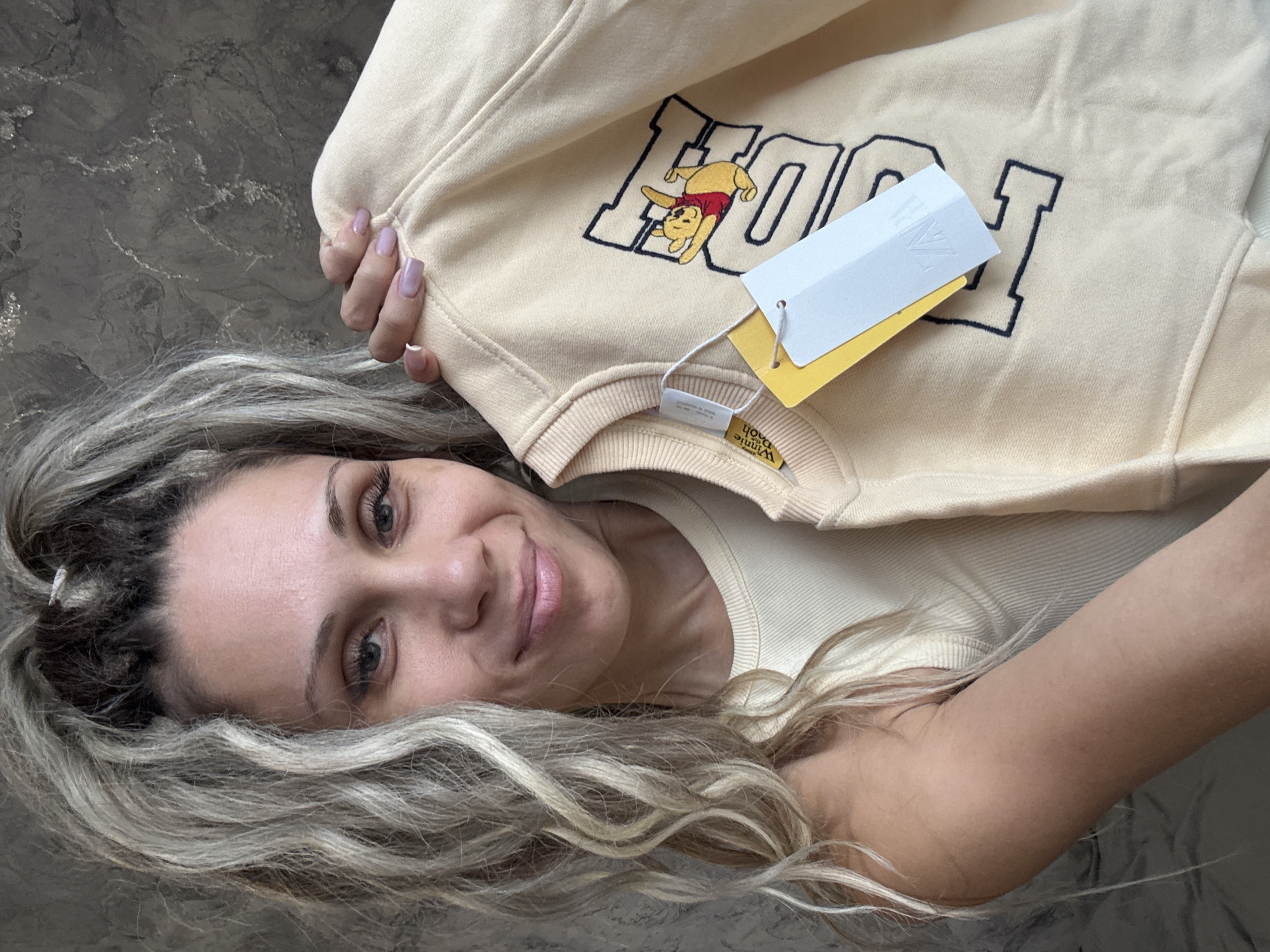 The image shows a woman holding a light yellow sweatshirt with a Winnie the Pooh graphic and the letters "POOH" in a collegiate font. She has blonde hair styled in dreadlocks and is wearing a cream-colored ribbed tank top. The background is a textured gray wall. Two clothing tags are attached to the sweatshirt; one is yellow and features a Winnie the Pooh graphic and "Winnie the Pooh" text, and the other is white with a "ZARA" logo. The woman is smiling and her fingernails are painted a light purple color. The scene appears to be indoors. There is no information in the image that indicates the location is in the city of Rusanovo, Russia.