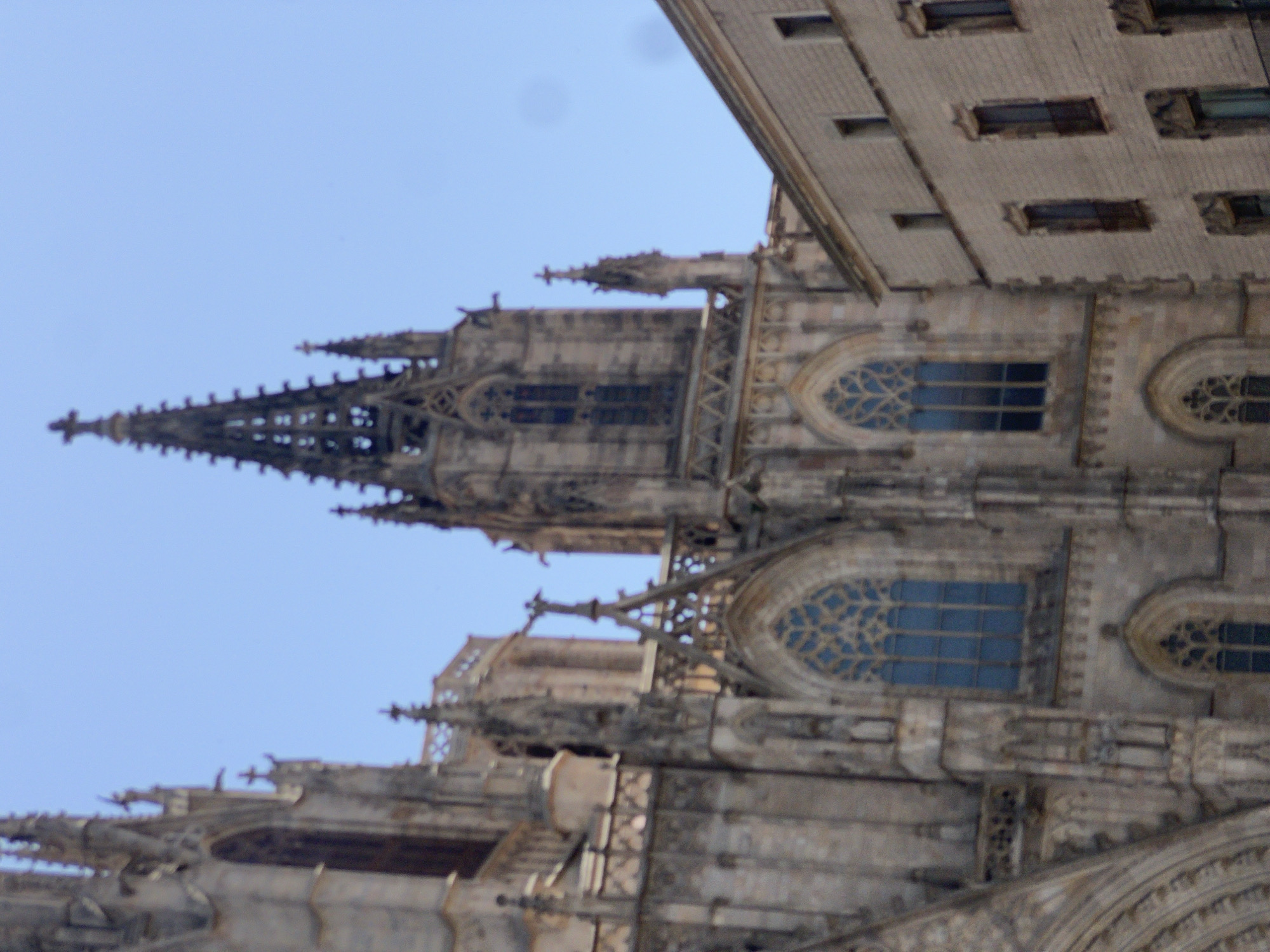 This media file captures a close-up, angled view of the intricate Gothic architecture of a large cathedral. The sky, a clear light blue, suggests it is daytime, likely morning or afternoon, with no visible clouds indicating fair weather. The image focuses on the detailed stonework, including spires, arched windows with stained glass, and decorative carvings. Several gargoyles are visible, characteristic of Gothic cathedrals, serving as both decorative and functional elements for water drainage. The sheer scale and complexity of the structure are emphasized by the close perspective, making the building appear immense.
The building's style suggests it is an older, historical landmark, possibly a cathedral or a similar religious or public building. The presence of pointed arches, ribbed vaults (implied by the overhead structures), and extensive ornamentation are hallmarks of Gothic architecture. The blue stained glass in the windows adds a touch of color to the otherwise stone-dominated facade. No people are visible in the frame, and the focus is entirely on the architectural details. The orientation of the image, tilted from the vertical, adds a dynamic perspective, drawing the viewer's eye upwards along the building's lines. The overall impression is one of grandeur, historical significance, and skilled craftsmanship. Given the location context of Barcelona, Spain, this structure is likely a notable landmark within the city.