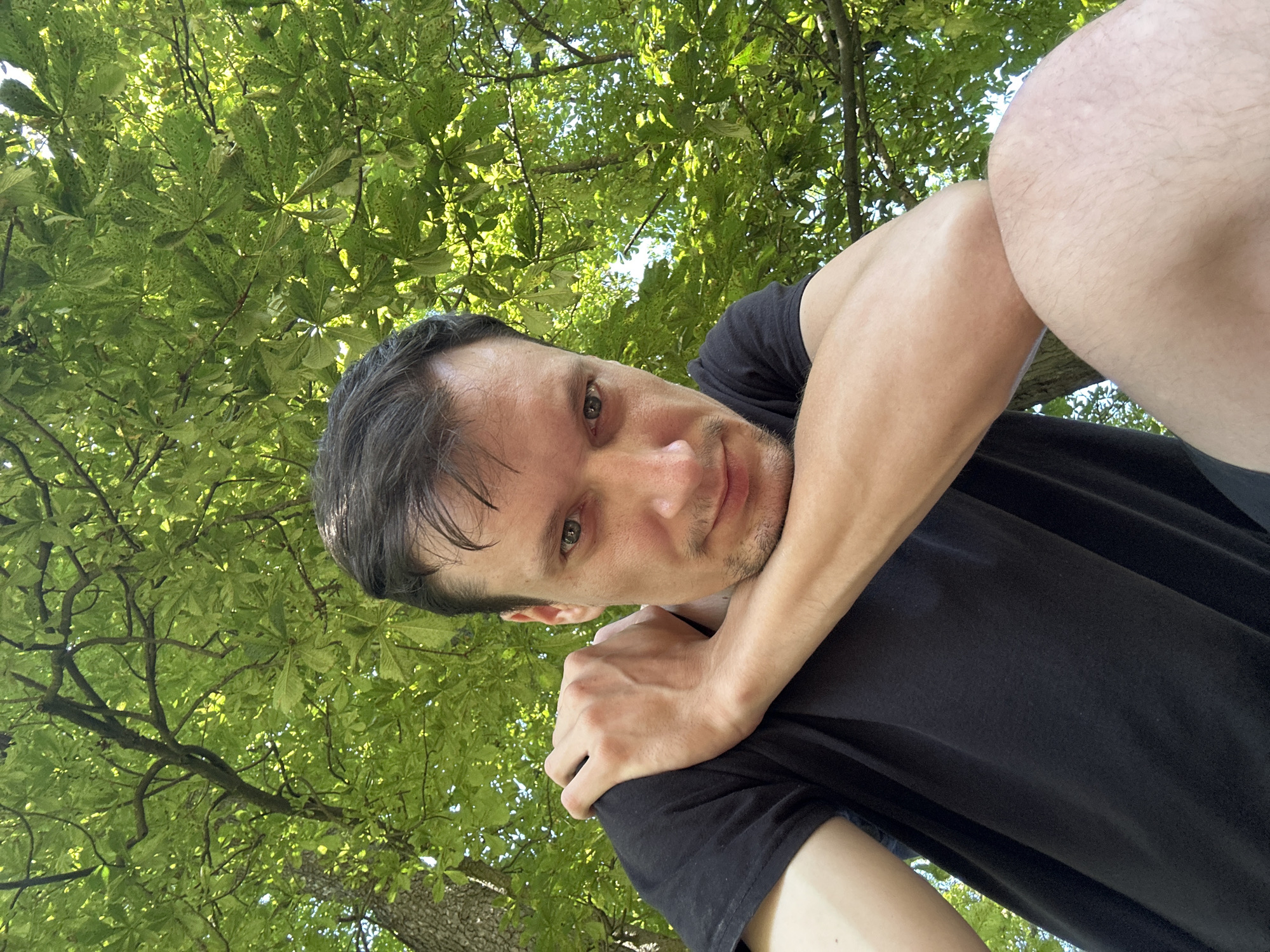 A low-angle selfie shows a man in a black t-shirt outdoors, looking towards the camera. He is holding his left arm across his chest, with his hand resting on his right shoulder. His right arm is bent, with his forearm resting on his thigh. The background is filled with lush green foliage, suggesting a park or natural setting. Sunlight filters through the leaves, creating dappled patterns on the man and the surrounding environment. The image appears to have been taken on a clear day. There is no visible indication of the city of Makiv, Ukraine, or any other specific urban context.