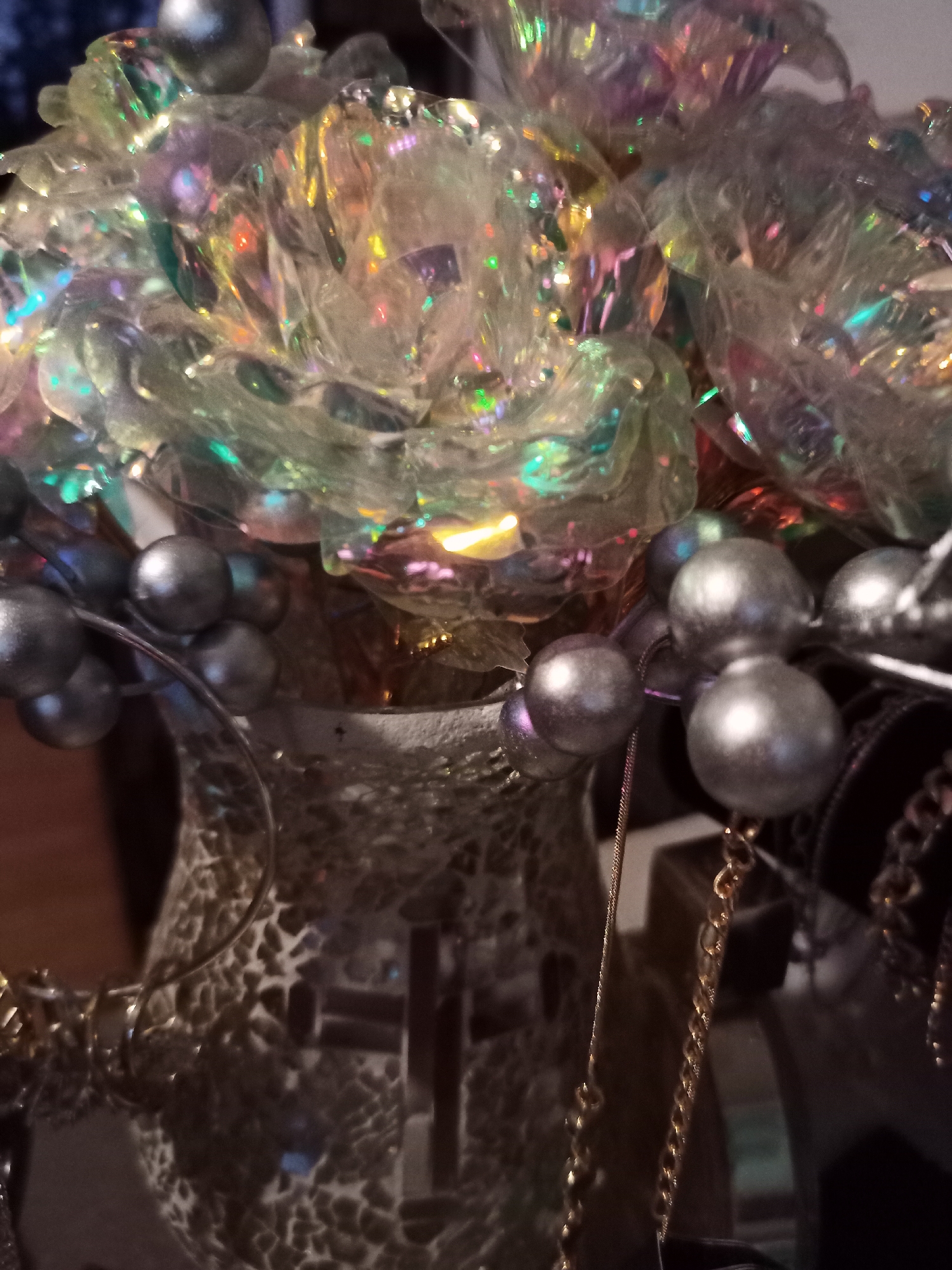 The image shows a close-up view of a vase filled with decorative, iridescent roses. The vase has a textured, mosaic-like pattern in shades of brown and cream. Several strands of metallic beads, some silver and some gold, are draped around the vase and the roses. In the background, other decorative items are visible, including chains and what appears to be a dark fabric. The lighting creates a reflective and somewhat abstract effect on the roses and beads. The image is a still life and does not depict any people, actions, or specific environment within Amarillo, United States of America.