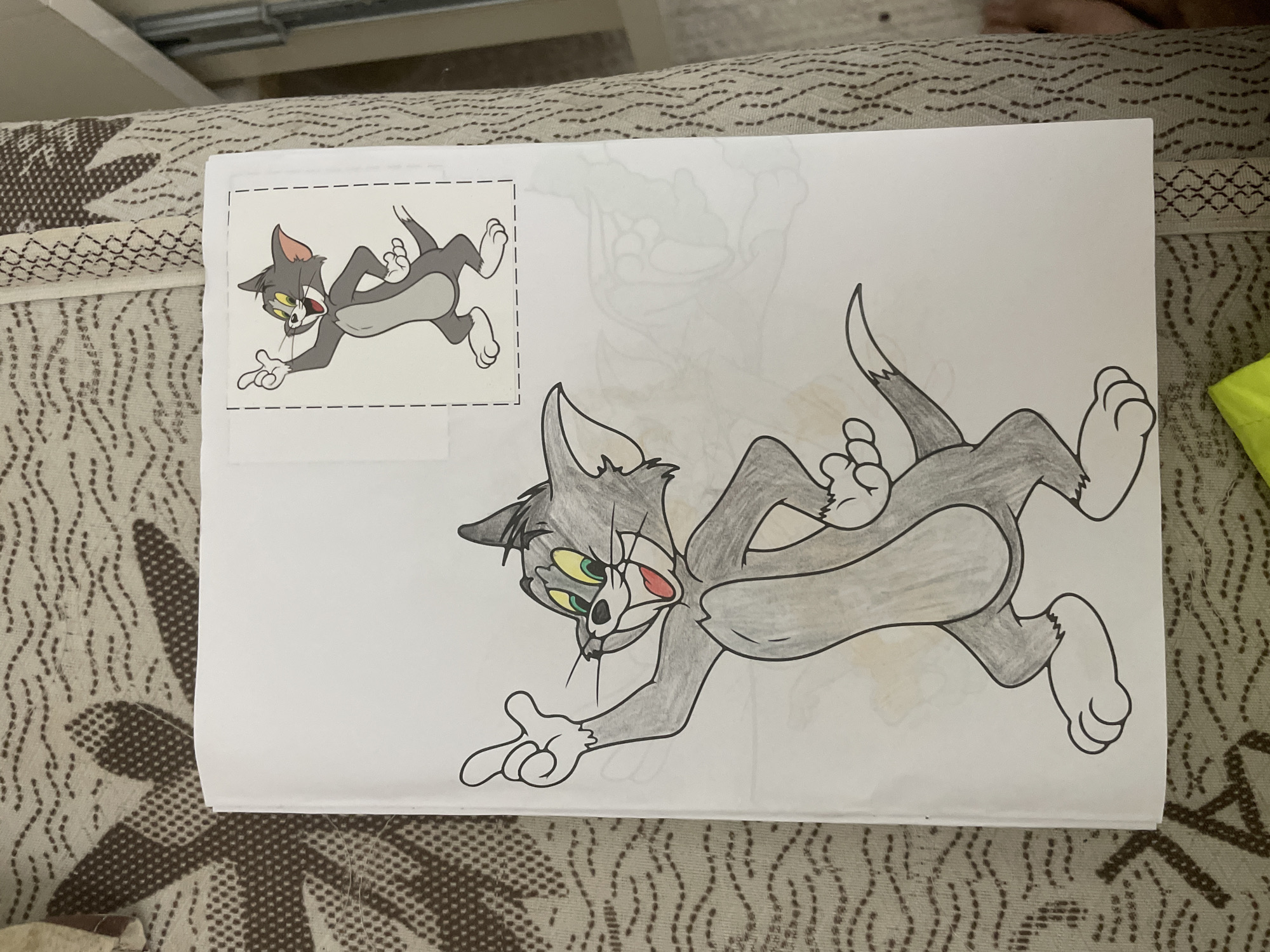 A coloring page featuring the cartoon character Tom from Tom and Jerry is depicted resting on a patterned fabric surface. The page shows two distinct illustrations of Tom. In the upper left corner, there is a smaller, fully colored illustration of Tom within a dashed-line border, suggesting it might be a reference image or a cutout. This Tom is shown in a dynamic pose, leaping forward with a surprised or excited expression.
Dominating the rest of the page is a larger, partially colored rendition of Tom, positioned at an angle. This larger Tom is also in a similar exaggerated, leaping pose, with wide yellow and green eyes, an open mouth showing a red tongue, and extended limbs. His fur is colored in shades of gray and black, with the inside of his ears and mouth colored red. White areas remain on his hands, feet, belly, and parts of his face, indicating the coloring process is incomplete. A faint, uncolored outline of another character or sketch is barely visible on the page to the right of the smaller Tom.
The paper lies on a textured surface, likely a couch or blanket, which features a repeating pattern of small dark dots on a light brown or beige background, interspersed with larger, stylized leaf-like motifs. A small portion of a bright lime green object is visible at the right edge of the frame. The overall scene suggests an indoor setting where a coloring activity is in progress. The image does not contain specific visual cues to definitively determine the location as Ihtiman, Bulgaria, beyond the general indoor setting. No text is visible in the image.