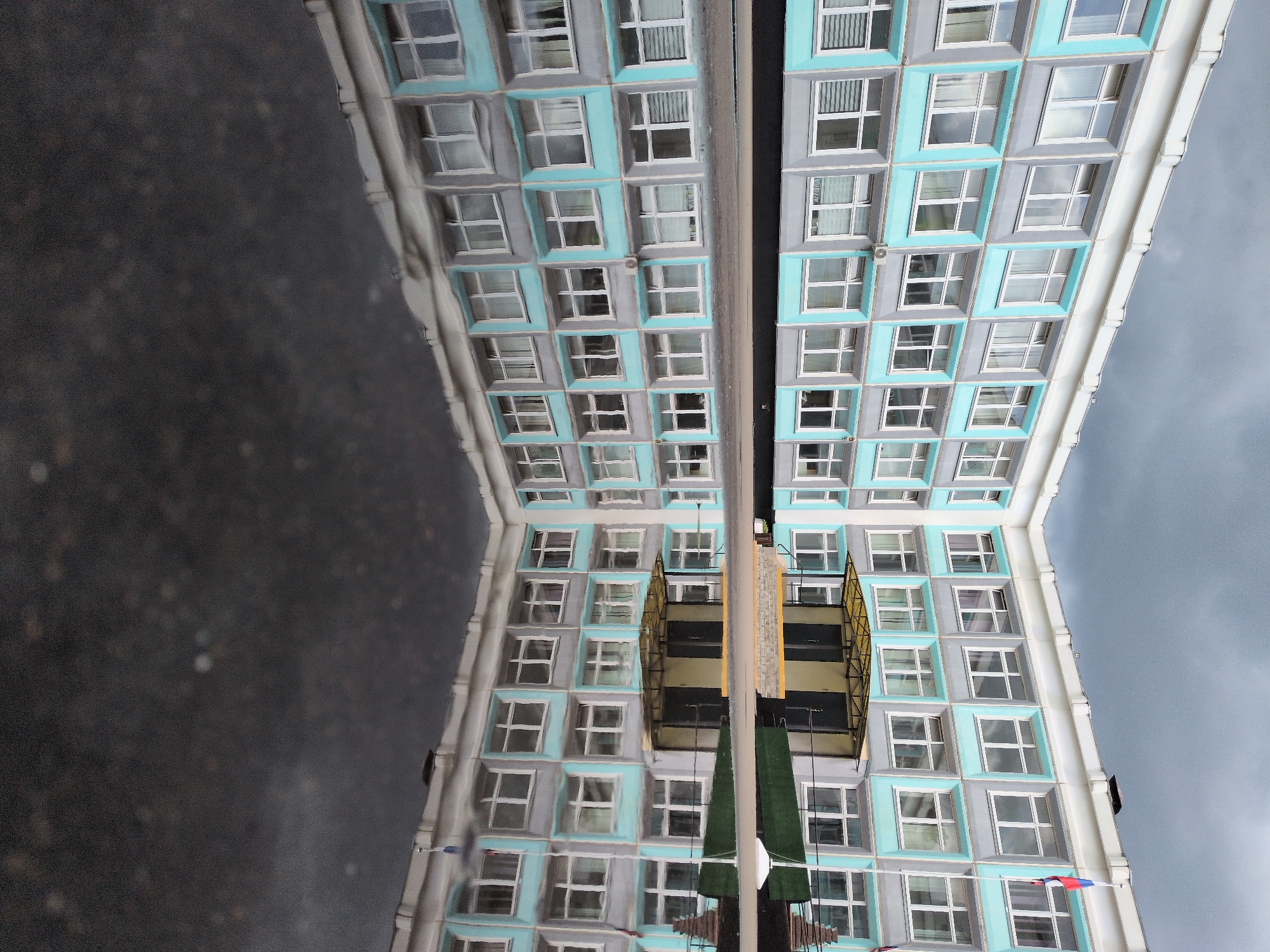 The image shows a reflection of a building with a modern facade in a puddle. The building has a grid-like pattern of windows framed in light blue and gray. A structure with a gold-colored lattice appears to be attached to the building. A dark pole extends from the structure into the reflected image. In the background, dark cloudy skies are visible, contrasting with the lighter colors of the building. The reflection indicates a wet surface, likely after rain. The presence of a Russian flag suggests the location is in Russia, and the architectural style is consistent with modern buildings found in Moscow.