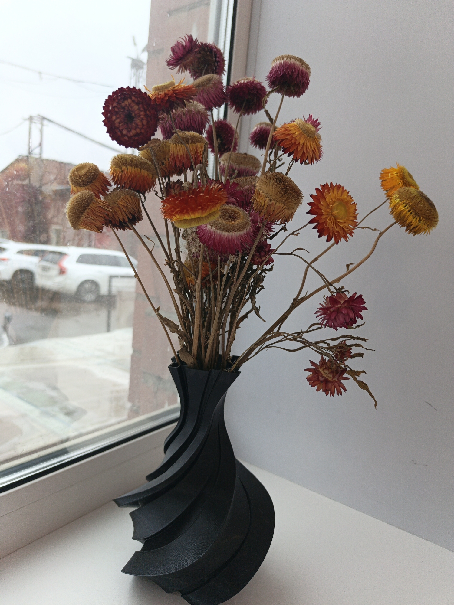 A still life image presents a black, artistically designed vase holding a bouquet of dried flowers. The flowers are of various colors including deep maroon, orange, and golden yellow. They are arranged with their dry stems protruding from the vase. The vase is placed on a white windowsill. Outside the window, the scene is blurry but appears to show a city street with cars and buildings, likely in Irkutsk, Russia.
