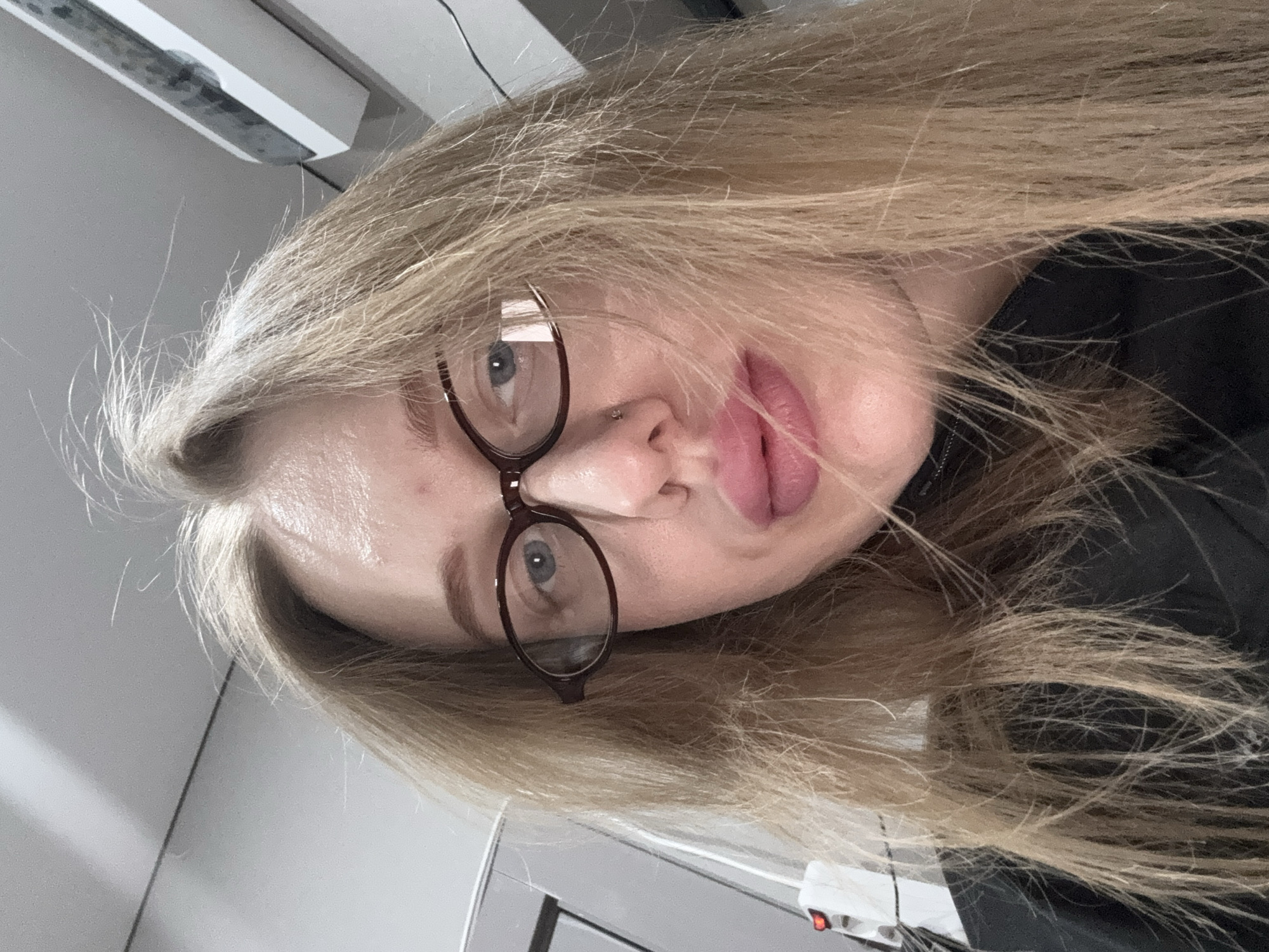 A selfie of a blonde woman wearing glasses. She is facing the camera at an unusual angle with her head tilted. Her lips are pursed. The background is an indoor setting that appears to be a workspace or office with visible lighting fixtures and cables. There is no visible information to establish the location is in the city of Azov, Russia.