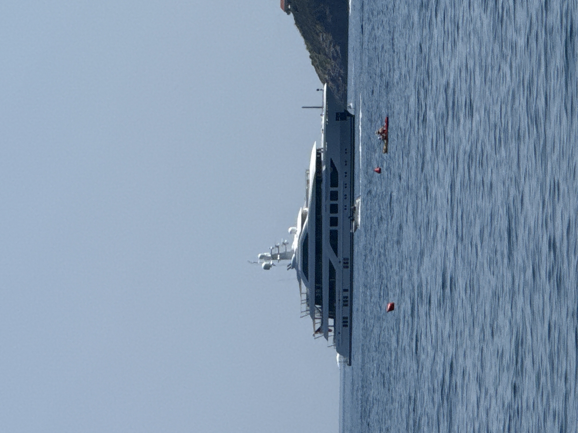 A large white yacht with multiple decks and a tall antenna is anchored in the blue water. To the right of the yacht, a green, tree-covered hill rises from the water, with a red-roofed building visible near the top. In the foreground, two people are in a red kayak, and a red buoy floats in the water. The sky is clear and blue.