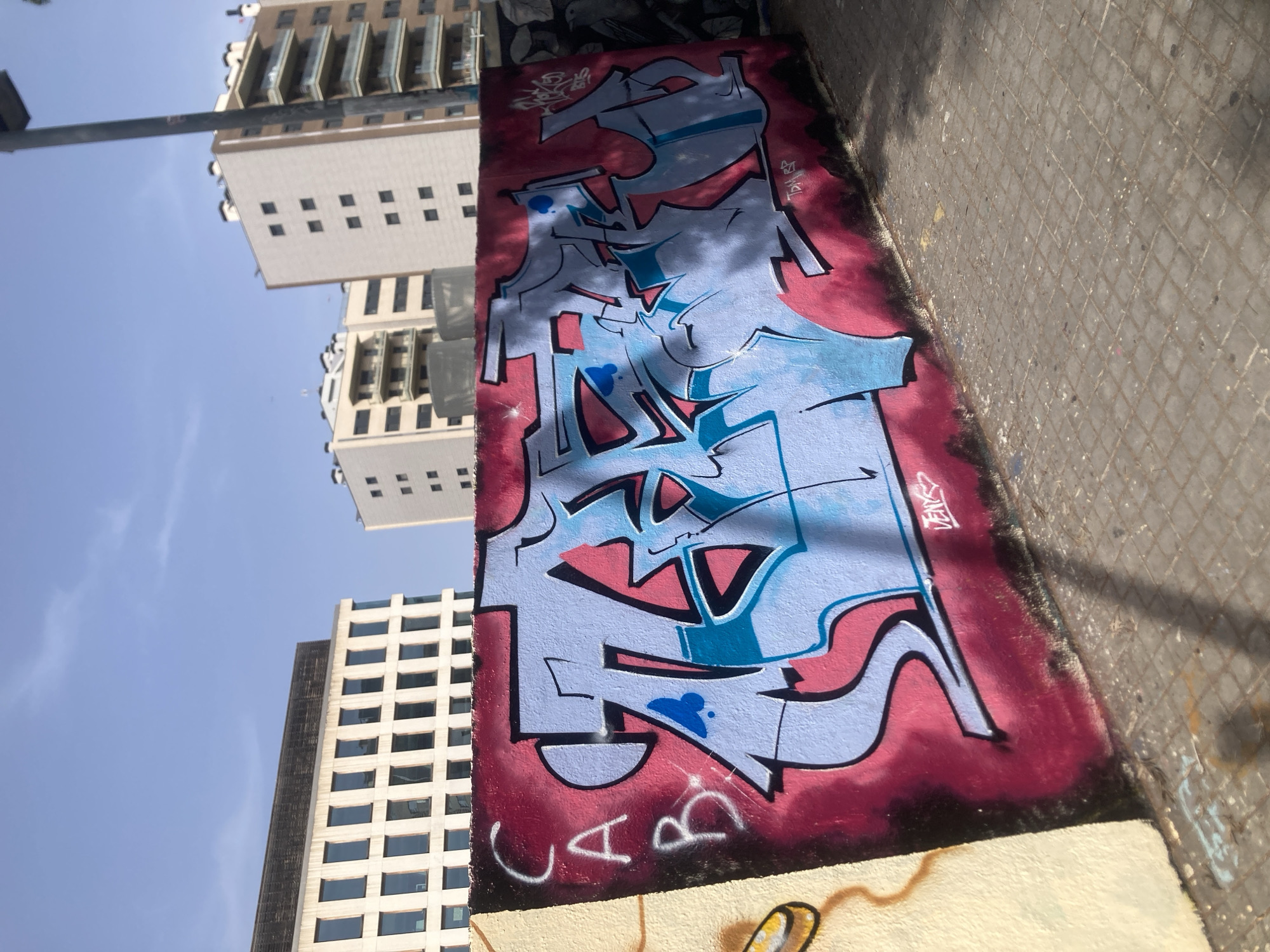 An outdoor, eye-level, full shot captures a graffiti-covered wall under a clear blue sky. The graffiti, predominantly in shades of light blue and pink, covers the majority of the wall's surface. On the left side of the wall, the letters "CAB" are written in white spray paint. The wall itself is set against a backdrop of modern, multi-story buildings. The ground in front of the wall is paved with square tiles. The scene appears to be in an urban environment, possibly a public space in Barcelona, Spain, judging by the architectural style of the buildings. There are no people visible in the image.