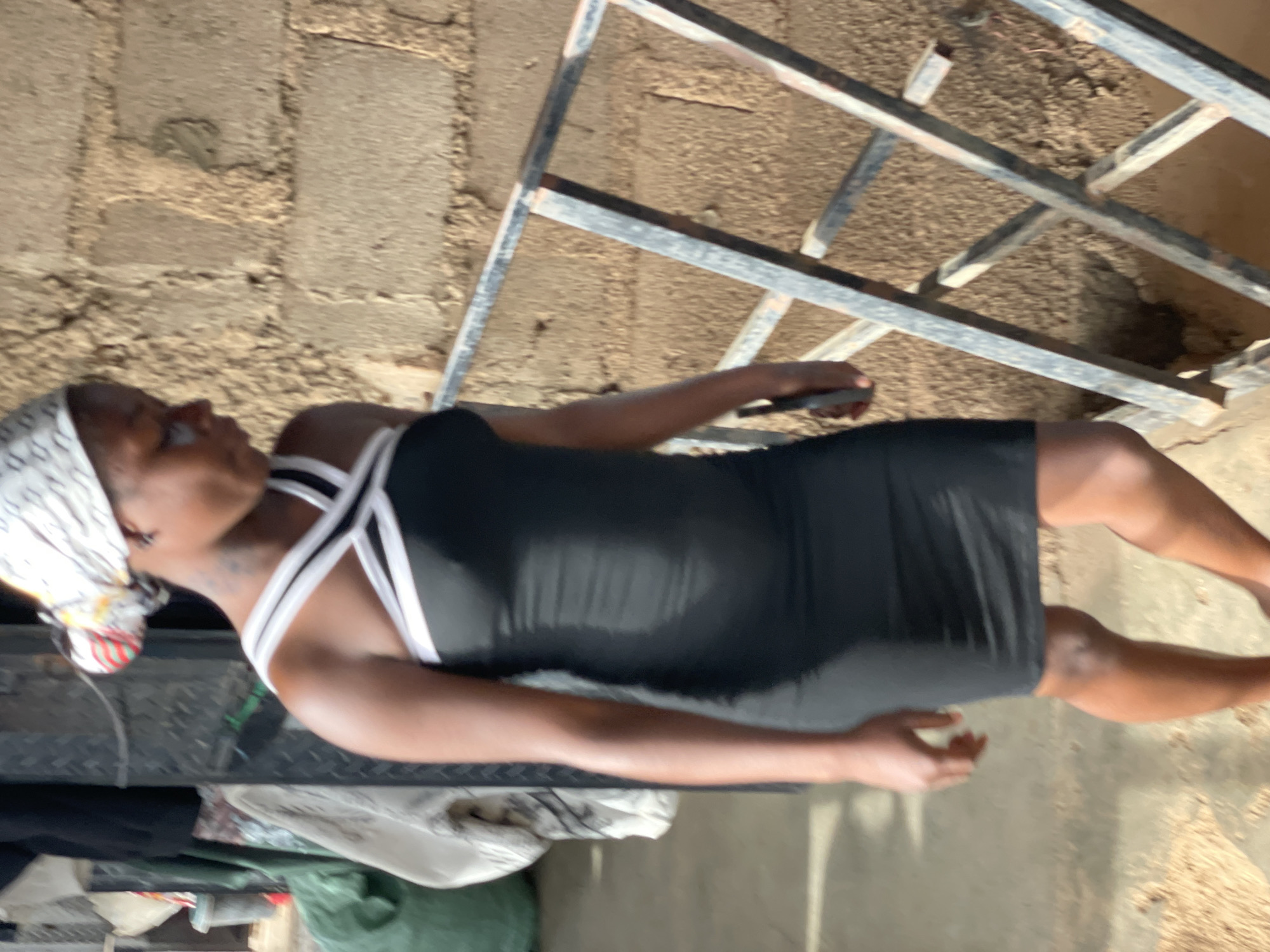 A person is standing in front of a brick wall and some metal bars. The person is wearing a black dress with white straps and a white headscarf. The person is facing to the left and their body is angled towards the viewer. The person's right arm is extended slightly and their hand is visible. The background appears to be a construction site or an area with rough terrain. The lighting suggests it might be daytime, but it's difficult to determine the exact time of day. There is no visible text in the image.