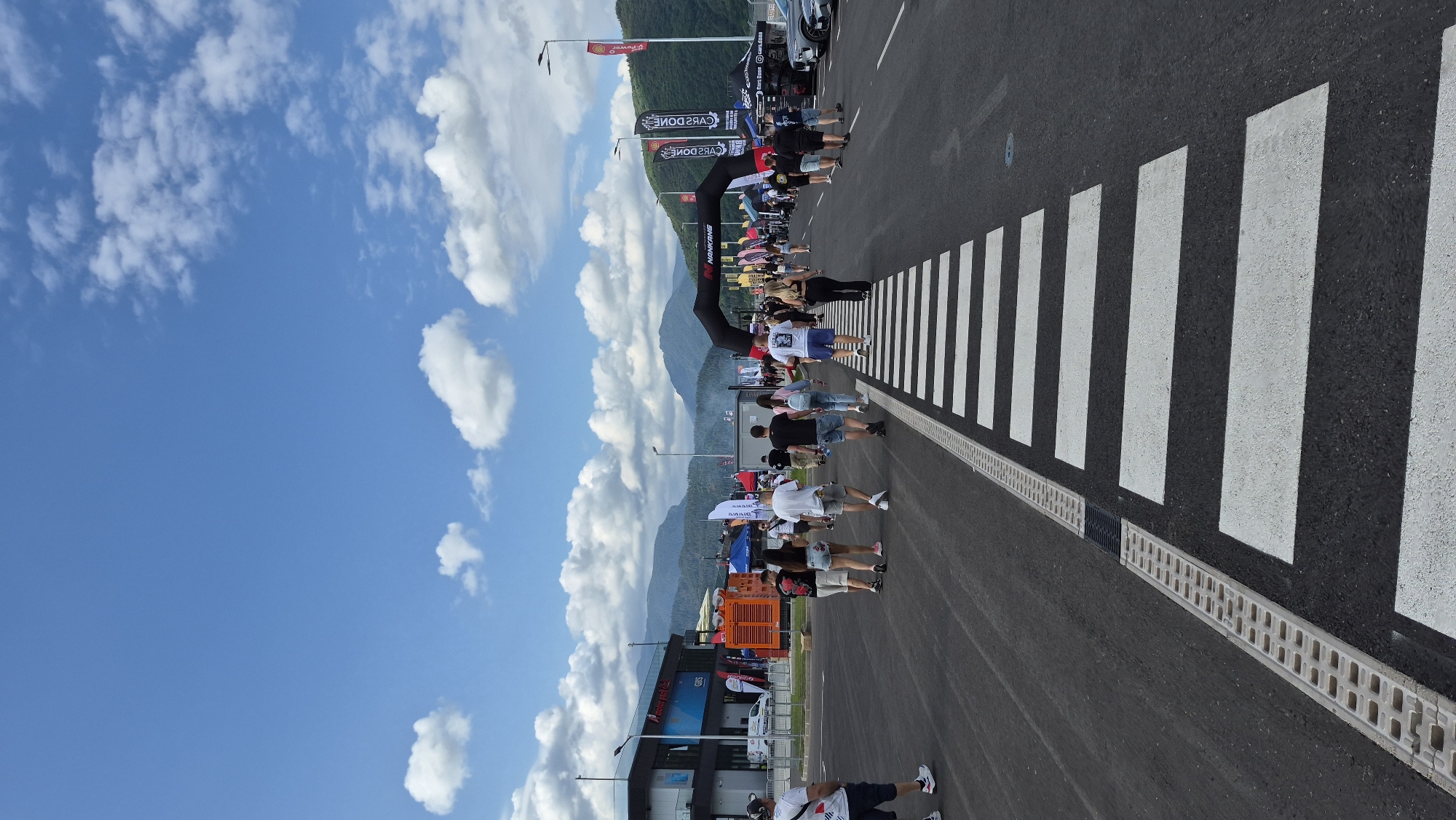 An outdoor daytime scene captures a street or parking lot filled with people walking towards an archway. The pavement is dark asphalt, marked with a prominent white pedestrian crossing. On the left, a modern building with large windows and signage is visible, along with an orange industrial generator. Tents and banners are set up throughout the area. In the background, lush green mountains rise under a bright blue sky dotted with fluffy white clouds. Several cars are parked on the right side of the image.