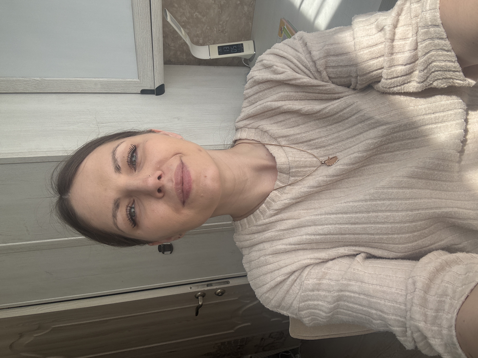The image shows a woman with fair skin, gray-green eyes, and dark hair wearing a light beige, horizontally ribbed sweater and a gold necklace with a pendant. Her expression is neutral, almost smiling. She is positioned in front of what appears to be a white desk or cabinet with a light wood grain finish. A small digital clock or device rests on the desk. Sunlight streams in from the upper right corner of the image.
There is no information about location in the city of Razdolie, Russia.