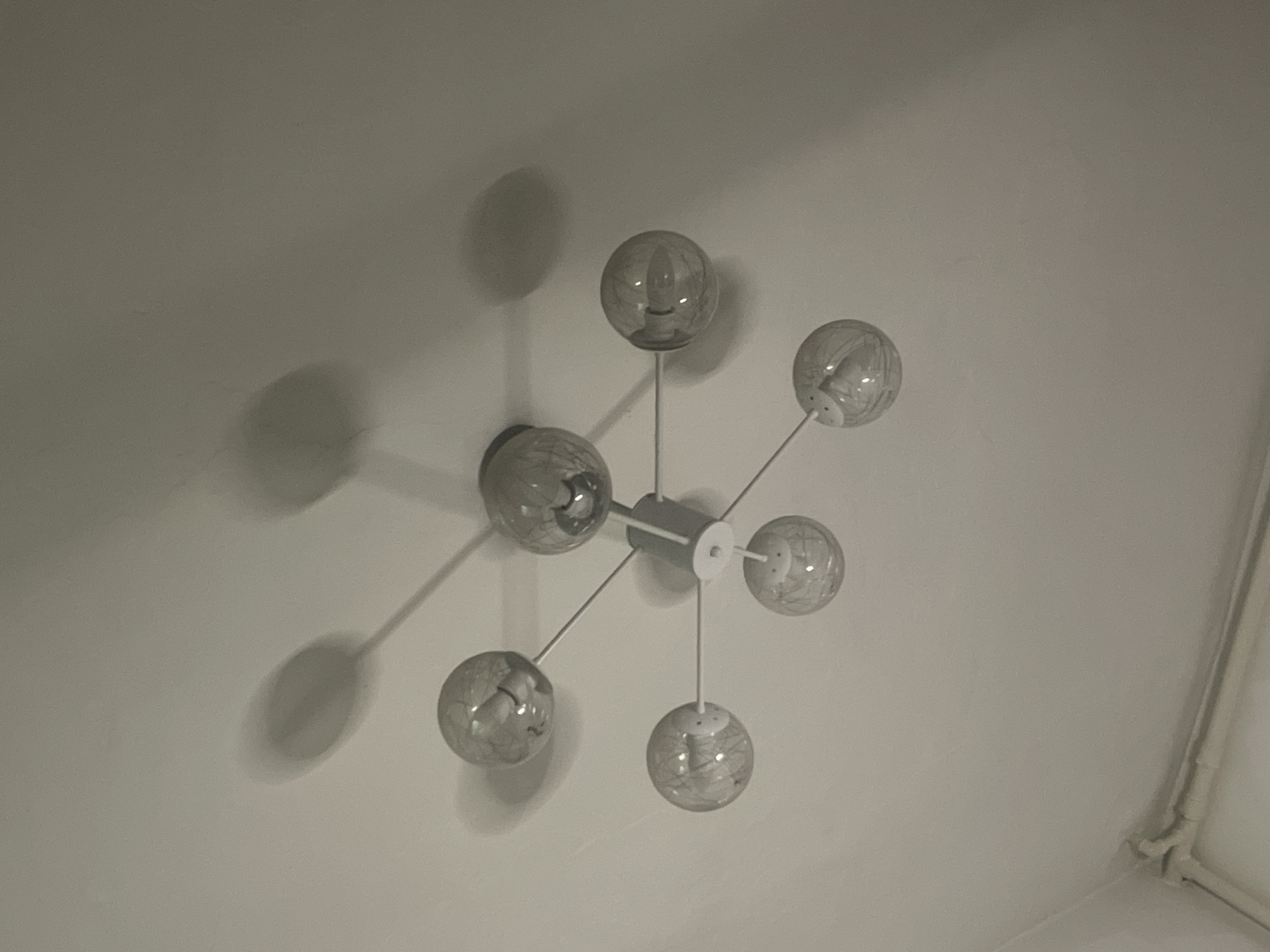 An indoor ceiling light fixture with a modern, atom-like design is captured from a low-angle perspective. The fixture has a central cylindrical element from which thin metal rods extend outwards, each terminating in a spherical glass shade. These shades are clear and appear to contain a decorative etched pattern. The bulbs within are visible, and some emit light, casting elongated, soft shadows onto the white ceiling. The ceiling itself shows a faint texture and a light source from above, likely from the fixture itself, illuminating the area. At the bottom of the image, part of a white pipe or conduit system is visible along the edge of the ceiling, suggesting it is a domestic or commercial interior space. The overall lighting is dim, emphasizing the shadows and the glowing bulbs. The image was taken in Bishkek City, Kyrgyzstan.
